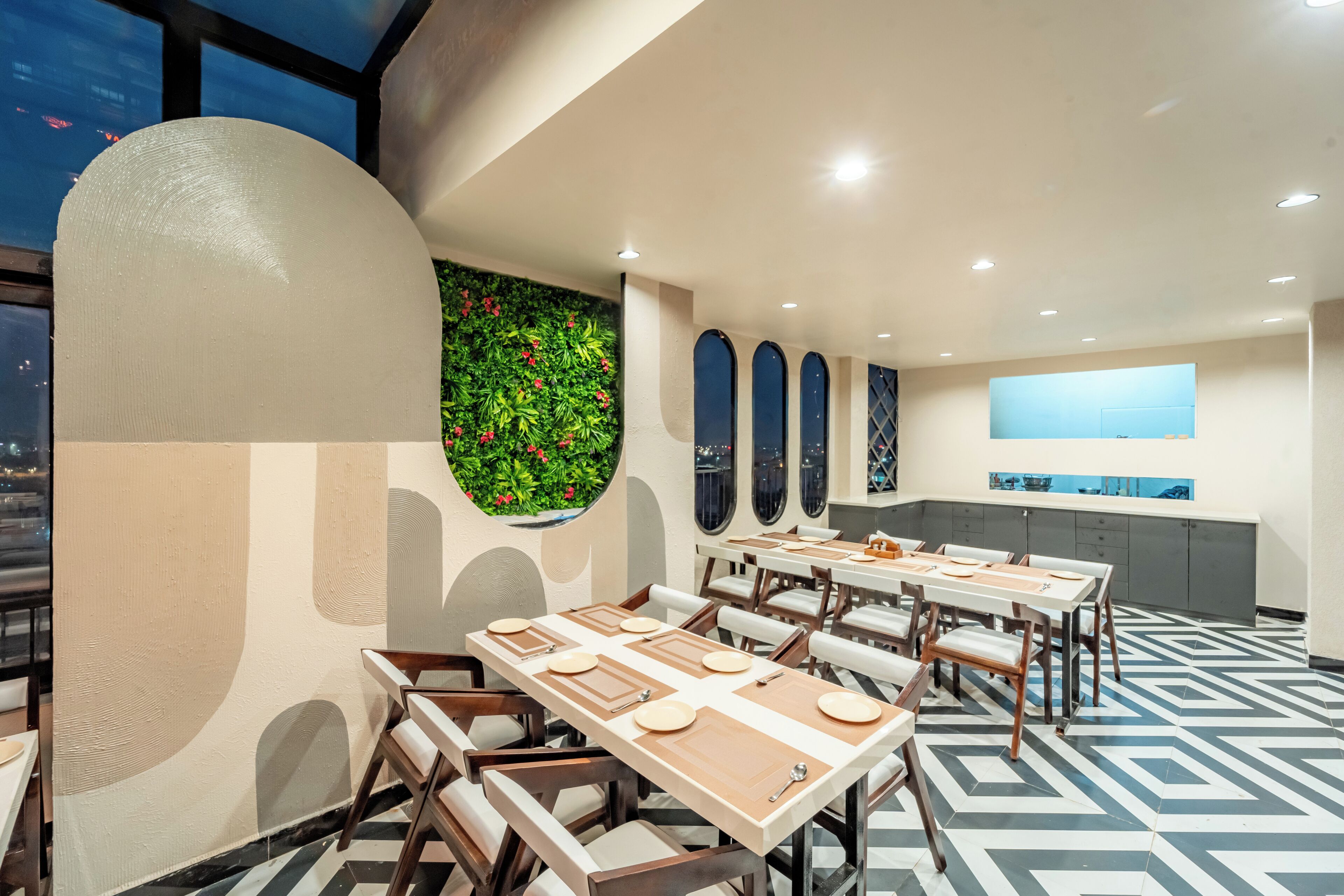 Un restaurant moderne avec un motif géométrique sur le sol, des tables rondes agrémentées de sets de table blancs et bruns, et un mur vert orné de fleurs rouges.