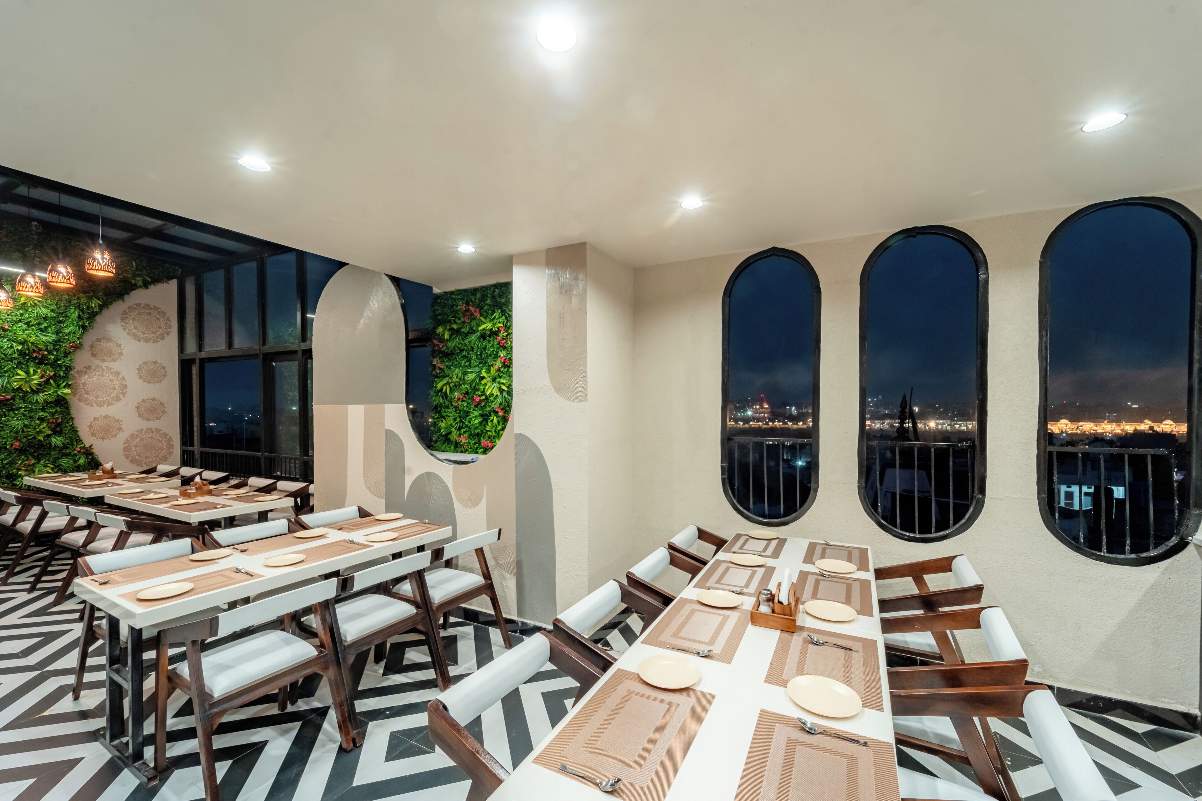 A restaurant with tables set for dining, large windows with city views, and a decorative wall with greenery.