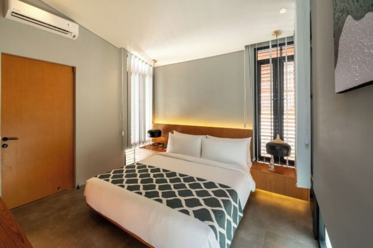 A hotel room with a bed, a nightstand, a lamp, a window with blinds, and an air conditioning unit.