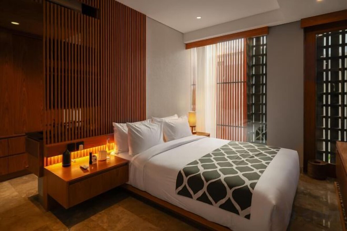 A hotel room with a bed, bedside table, lamp, and a view of the outside through a window.