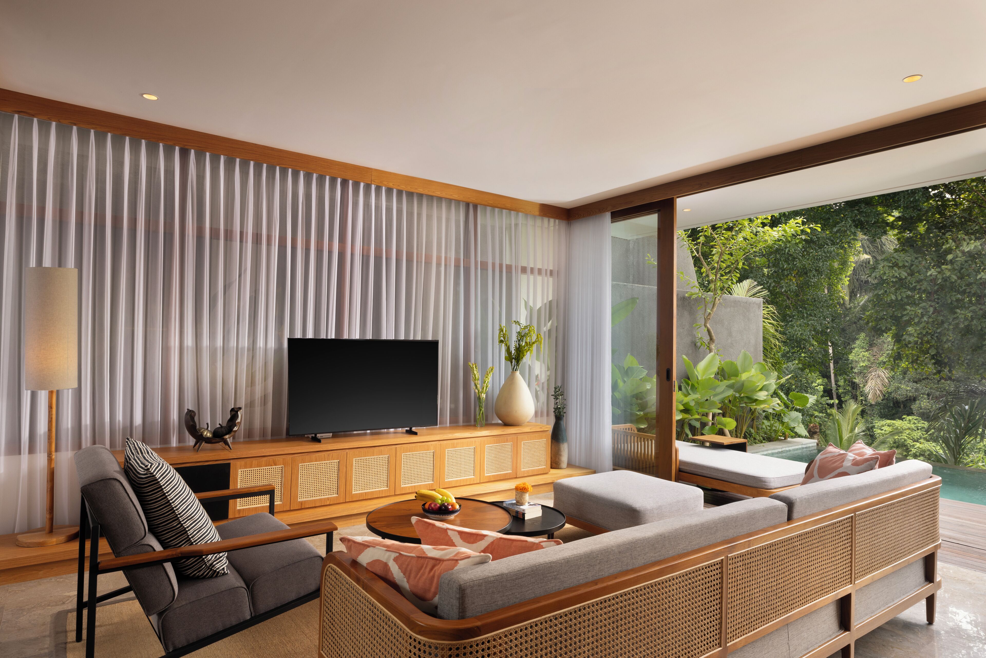 A modern living room with a flat-screen TV, a sofa, a coffee table, and a large window overlooking a lush garden.