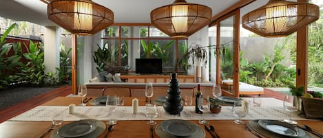 Un coin repas avec une table en bois dressée pour six personnes, entouré de grandes fenêtres offrant une vue sur une végétation luxuriante.