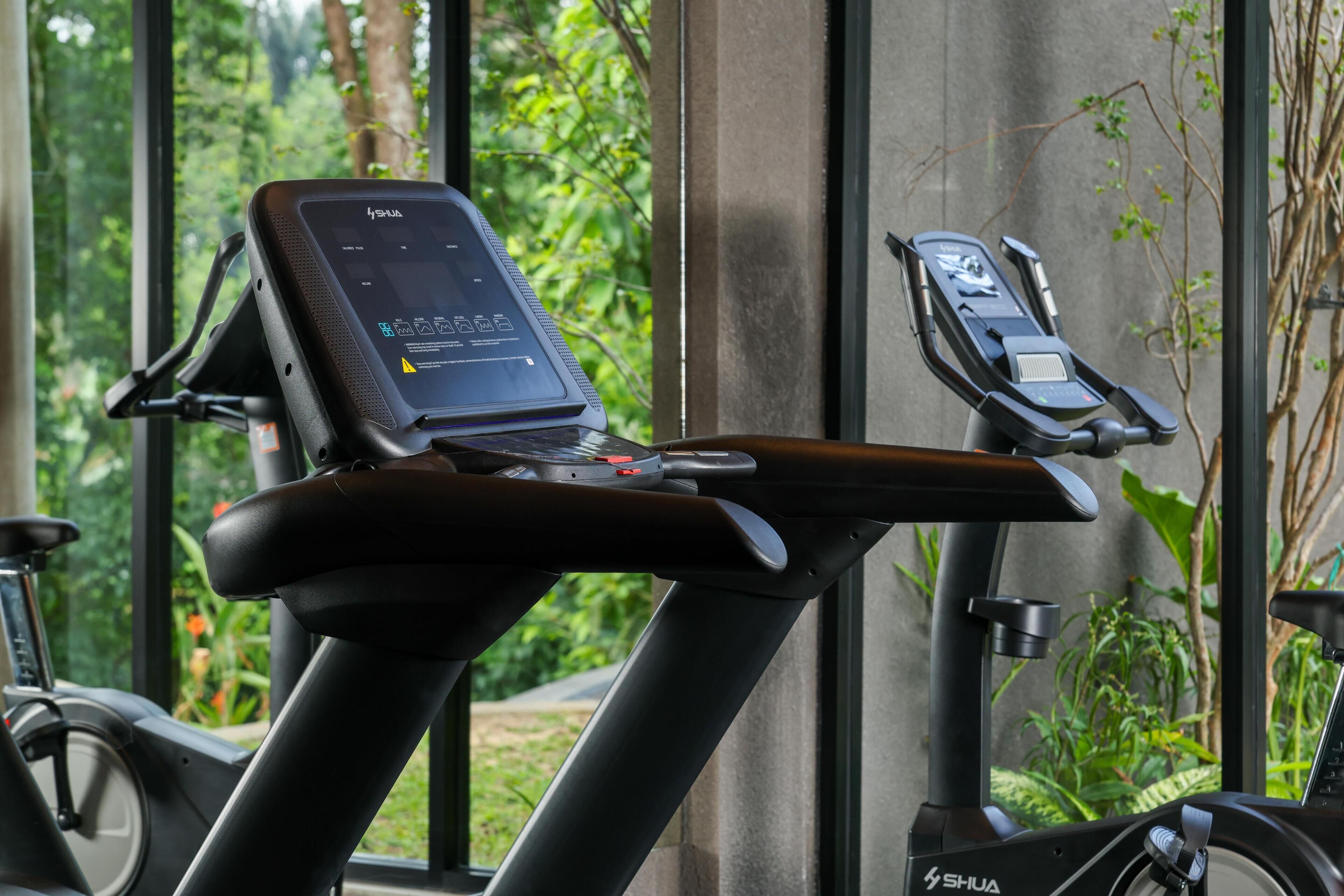 Una bicicleta estática con pantalla digital y pedal para los pies, ubicada cerca de una ventana con vista a follaje.