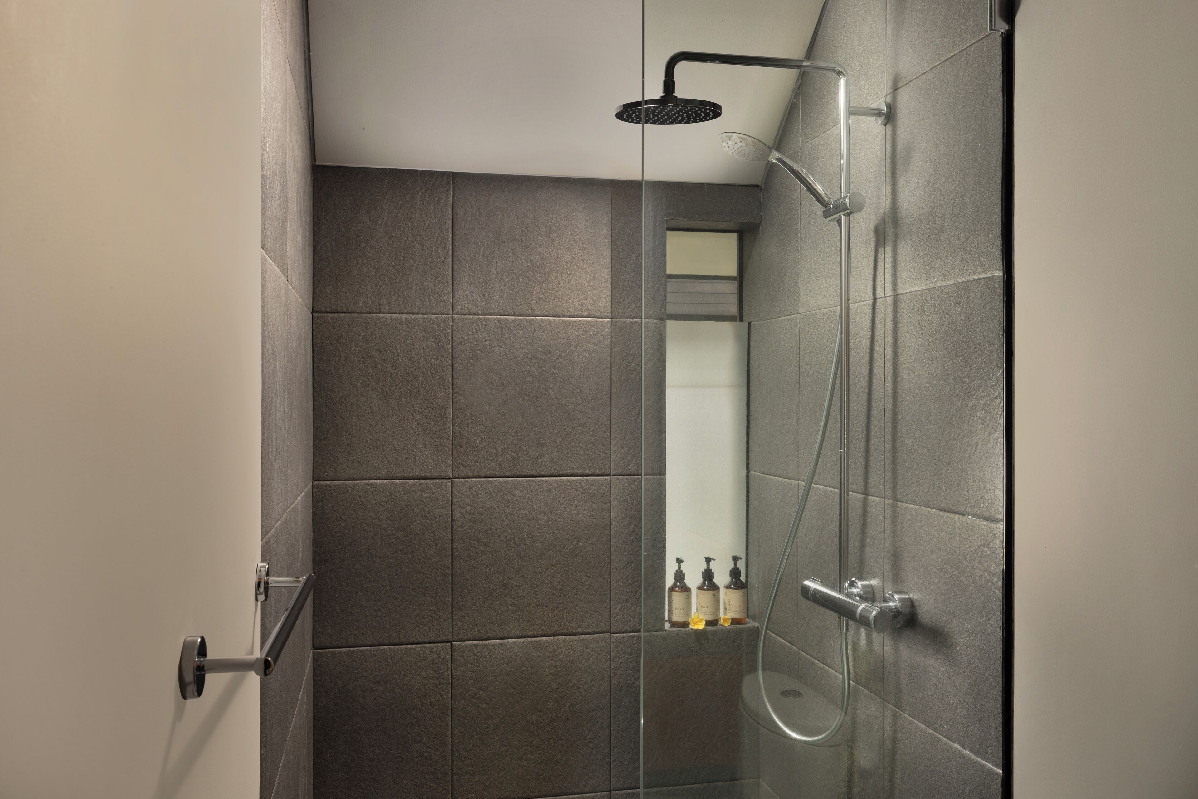 Un área de ducha moderna con puerta de vidrio, azulejos grises y un cabezal de ducha de mano.