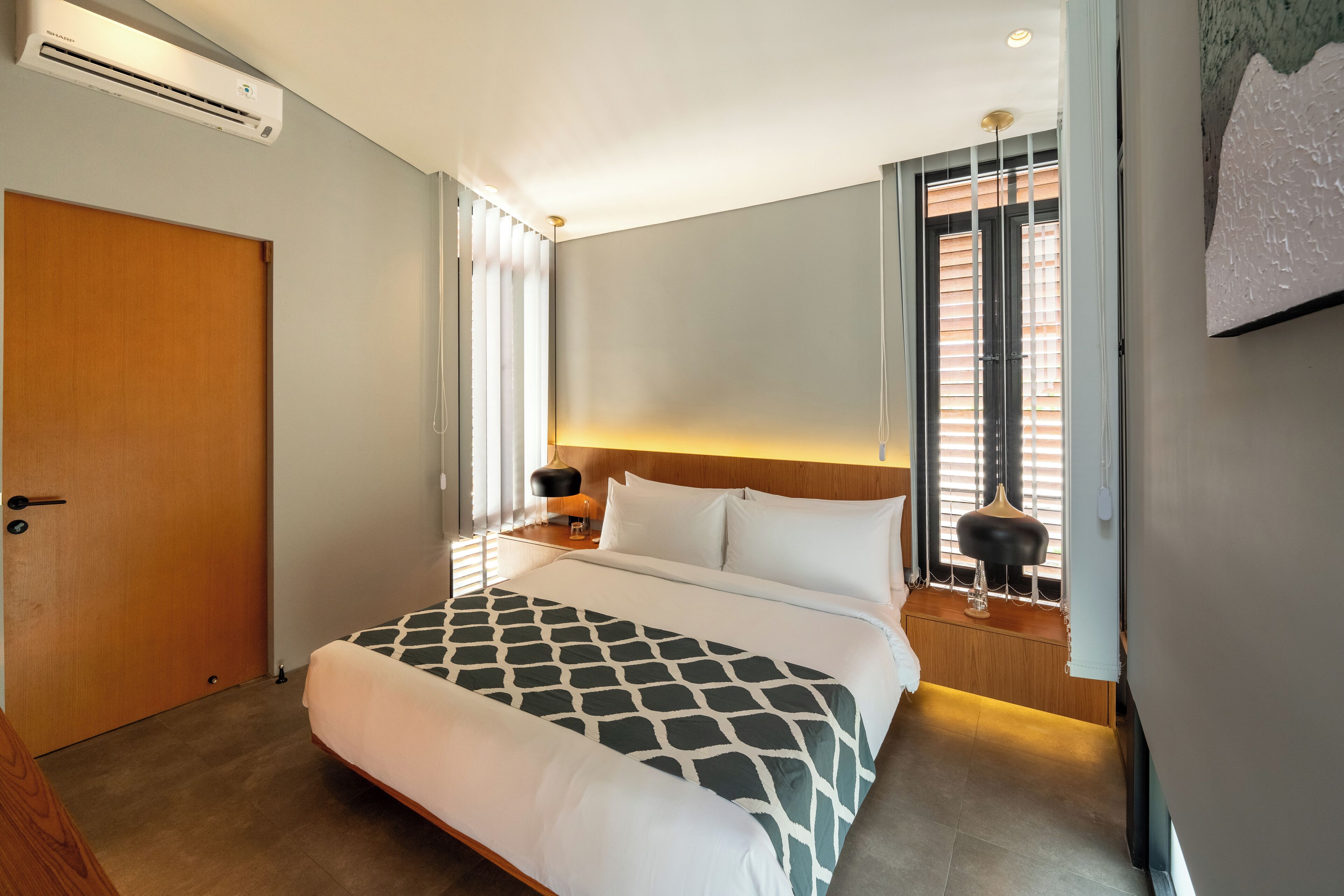 Hotellihuone, jossa on suuri sänky, yöpöytä lampulla, ikkuna, jossa on sälekaihtimet, ja ilmastointilaite seinällä.