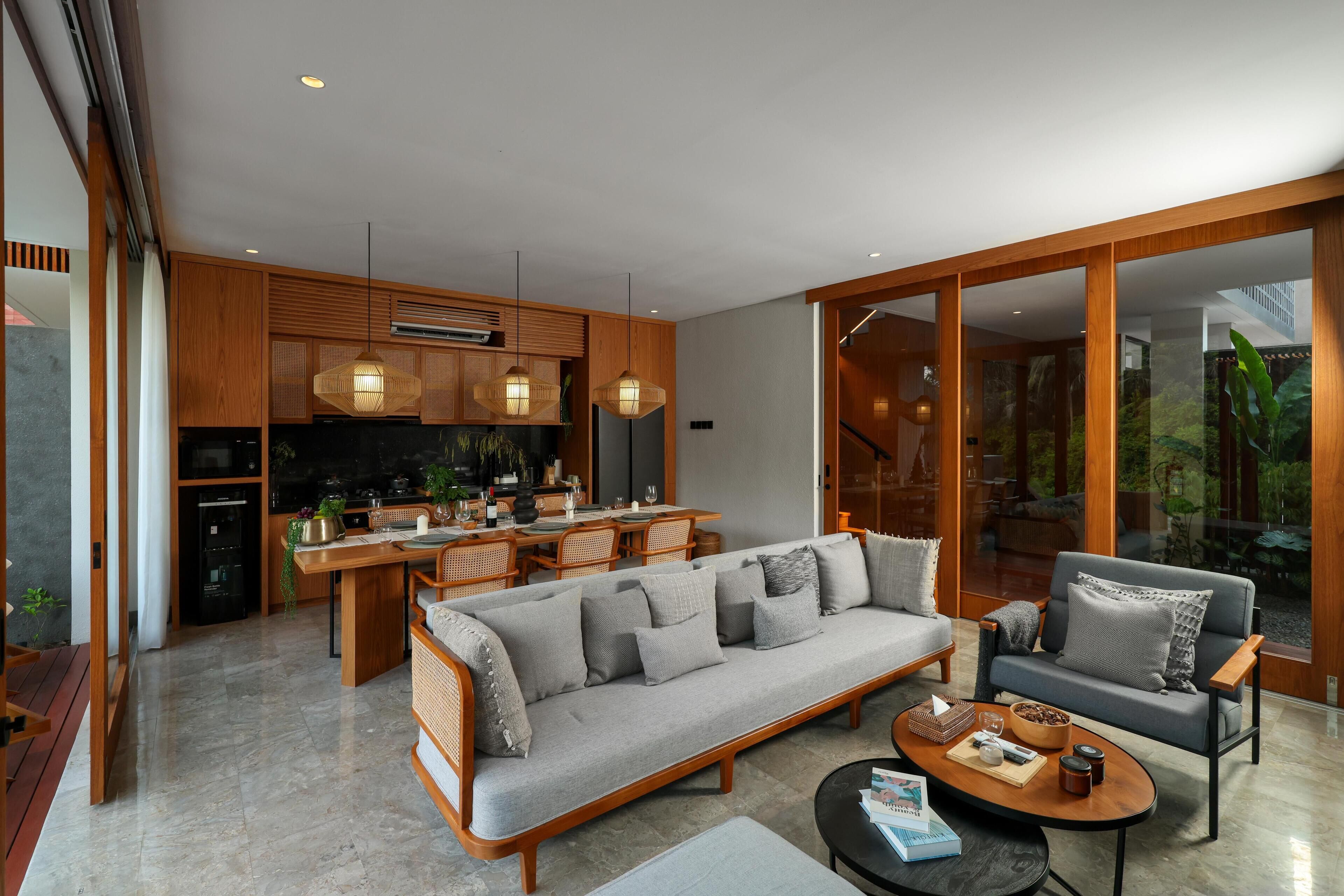 Ein modernes Wohnzimmer mit Sofa, Sessel und Couchtisch, angrenzend an eine Küche mit Holdekor-Schränken und Pendelleuchten.