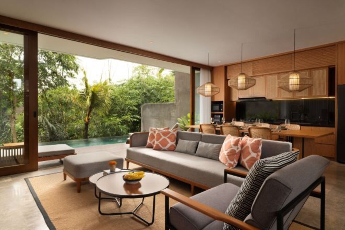A modern living room with a sofa, armchairs, a coffee table, and a TV. There is a dining area with a table and chairs. The room has large windows with a view of greenery outside.