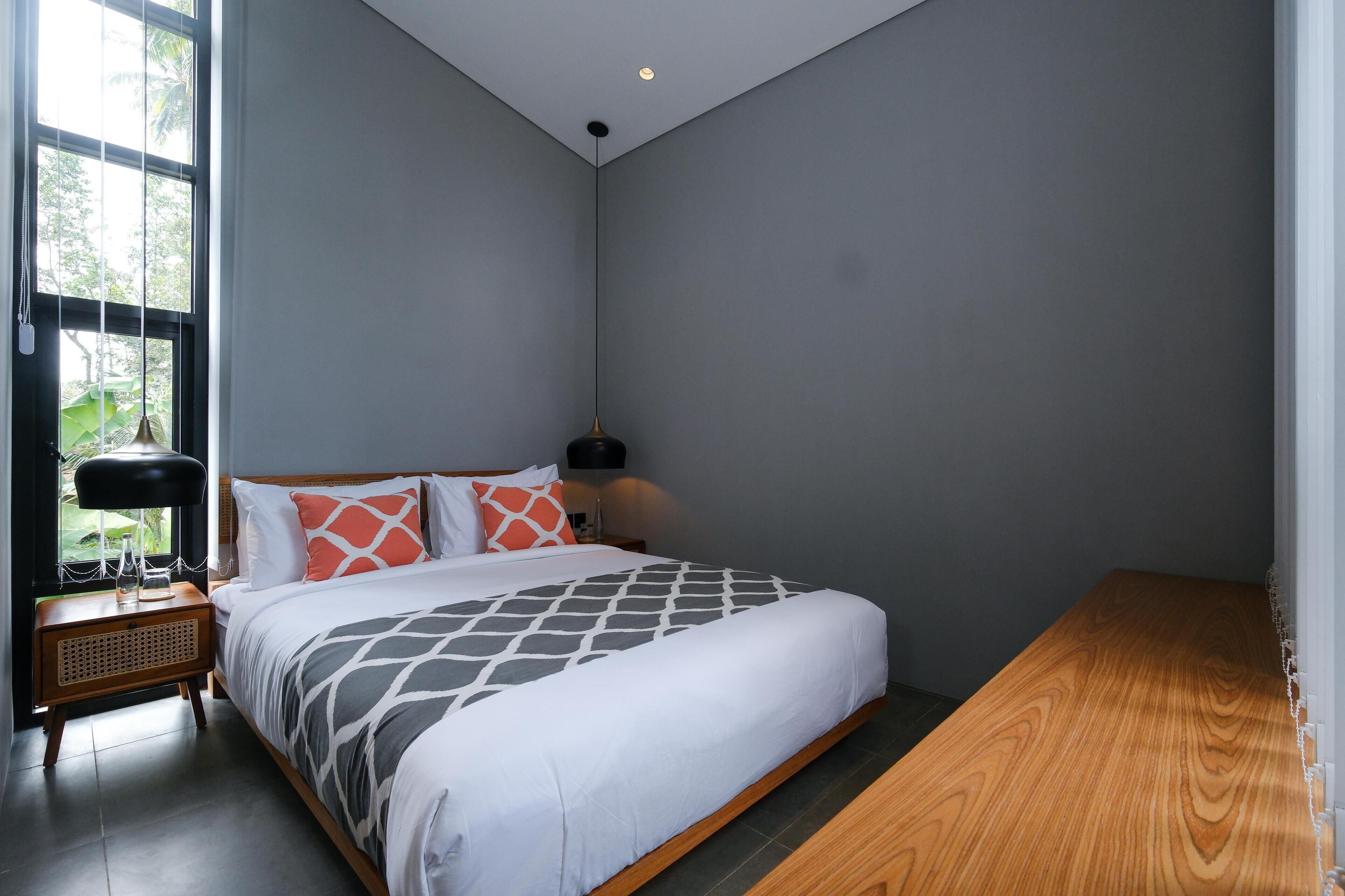 A modern bedroom with a large bed, a bedside table, a lamp, and a view of greenery outside.