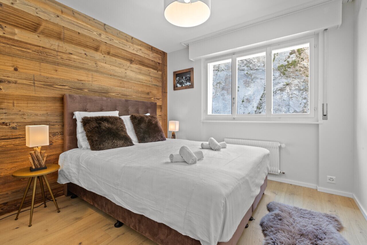 Ein Schlafzimmer mit einem Holzkopfende, weißer Bettwäsche, einem Nachttisch mit Lampe und einem Fenster mit Blick auf Bäume.