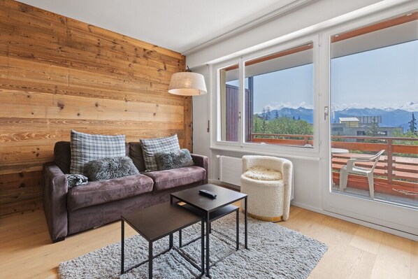 A modern living room with a wooden wall, a purple sofa, a small table, and a view of mountains through large windows.