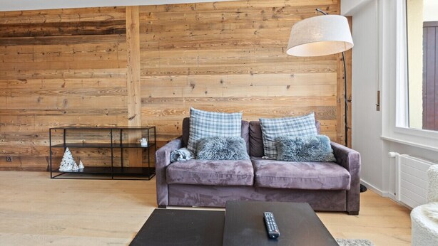 A living room with a wooden wall, a purple sofa with plaid pillows, a black coffee table, and a floor lamp. - Appartement Rénové, Calme et Cozy (Crans-Montana)