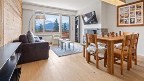 A modern living room with a sofa, dining table, and chairs, a TV mounted on the wall, and a large window with a mountain view. - Appartement Rénové, Calme et Cozy (Crans-Montana)