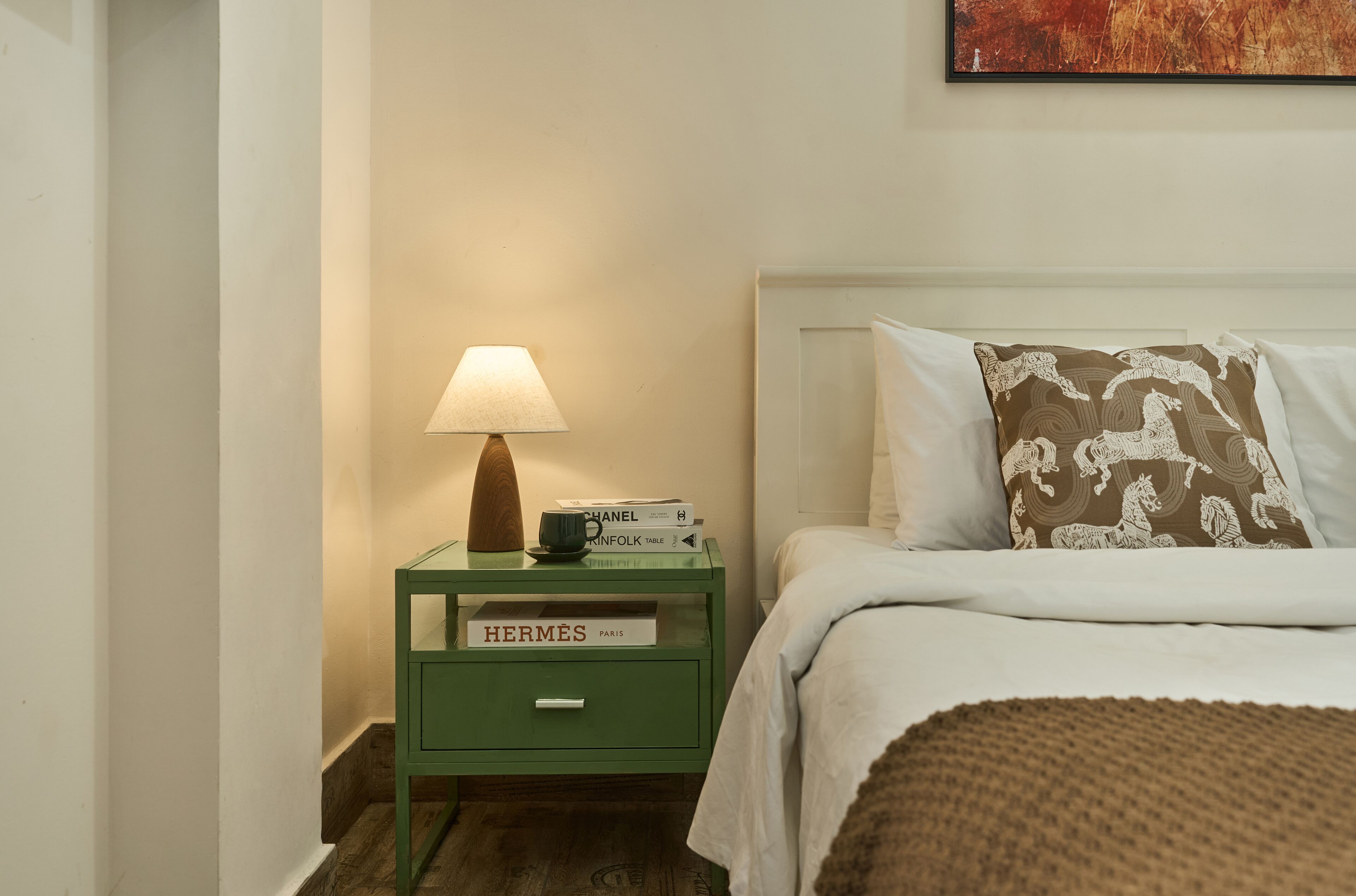 A neatly made bed with a pillow, a bedside table with a lamp, and a book titled 'HERMES PARIS' on the table.