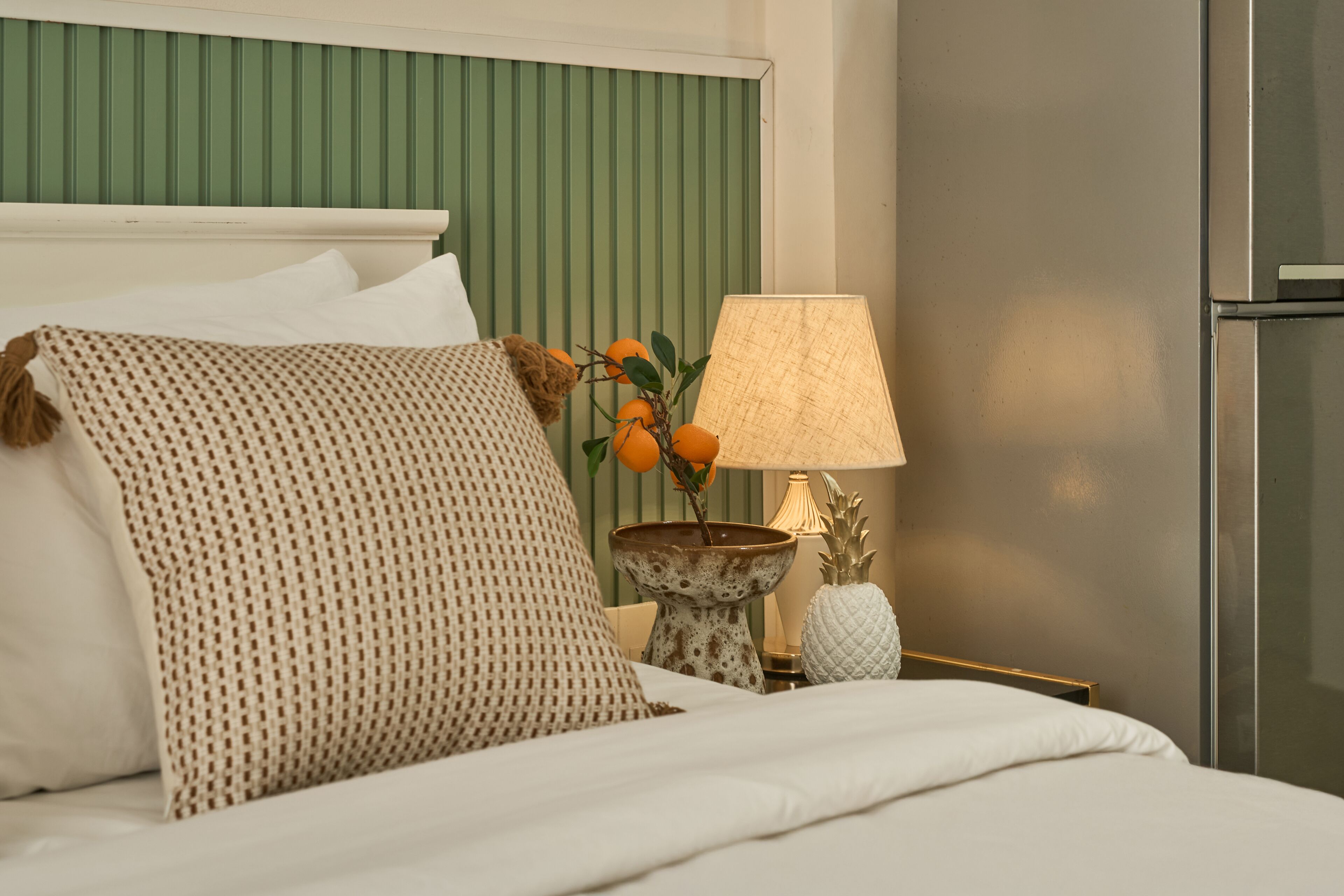 Una cama bien hecha con un cojín estampado, una mesita con una lámpara y una piña decorativa.