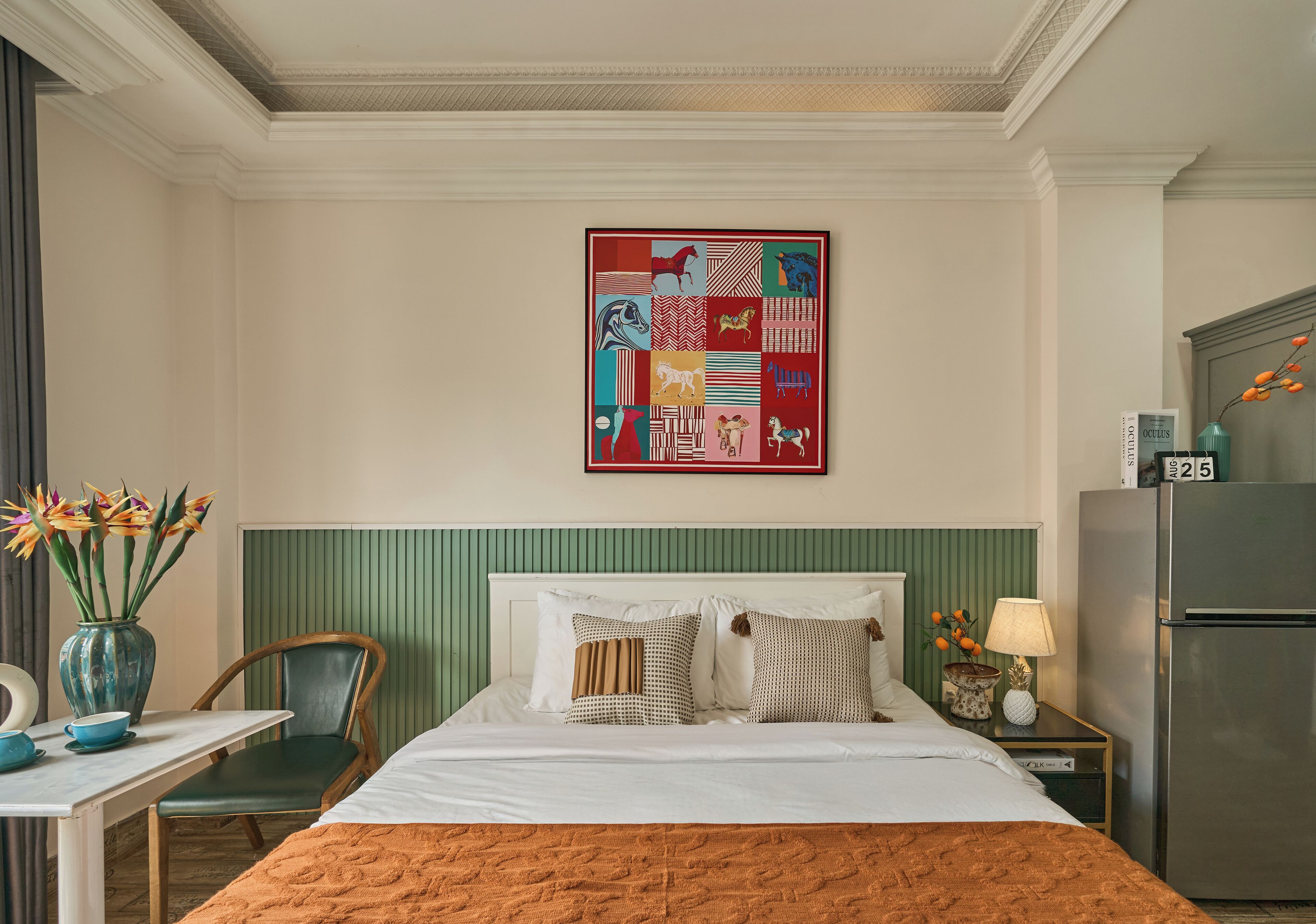 A hotel room with a bed, a chair, a table, a vase with flowers, a lamp, and a refrigerator.