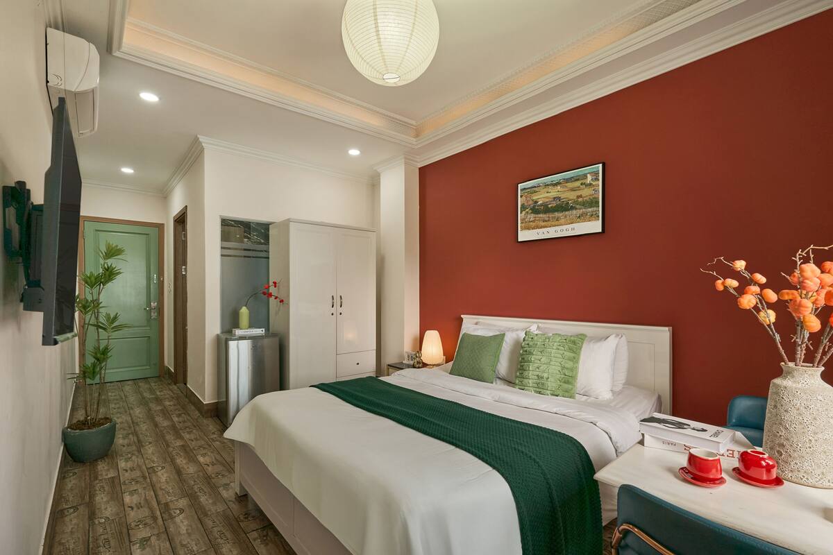 A hotel room with a bed, a desk with a lamp, a vase with flowers, and a painting on the wall.