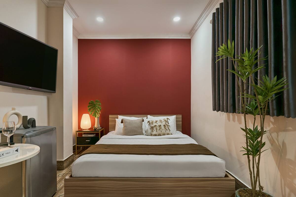 A modern hotel room with a red accent wall, a large bed with white bedding, a flat-screen TV mounted on the wall, a small desk with a lamp, and a potted plant.