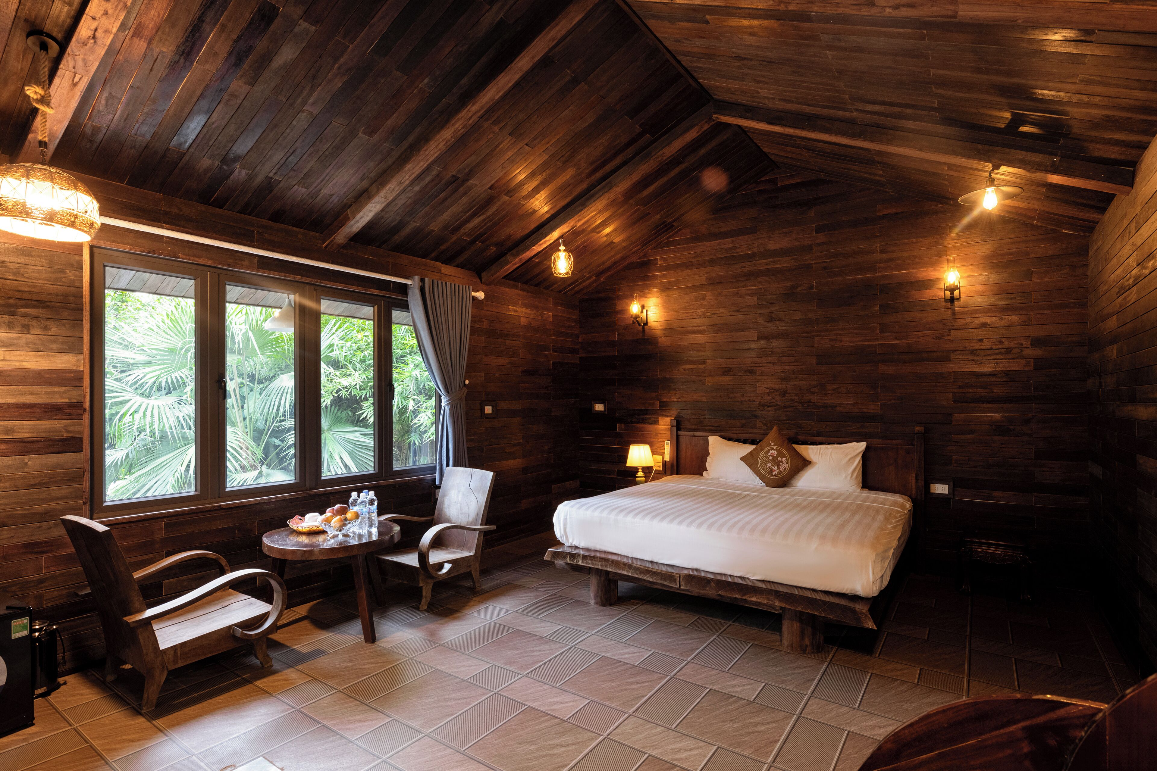 A wooden room with a bed, a chair, a small table with food, and a window with curtains.