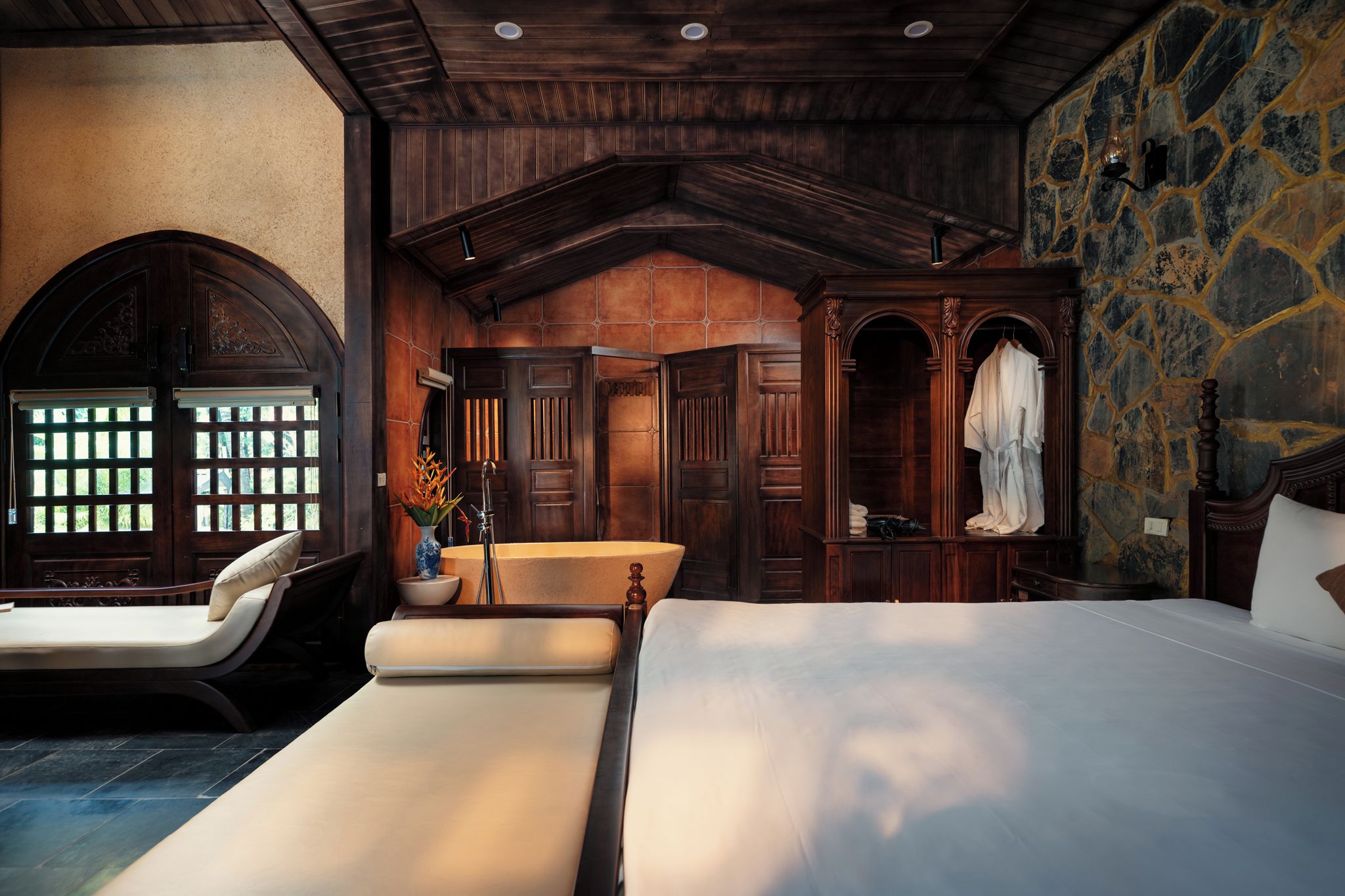 A hotel room with a large bed, a sitting area with a chair, a vanity with a mirror, and a stone wall.
