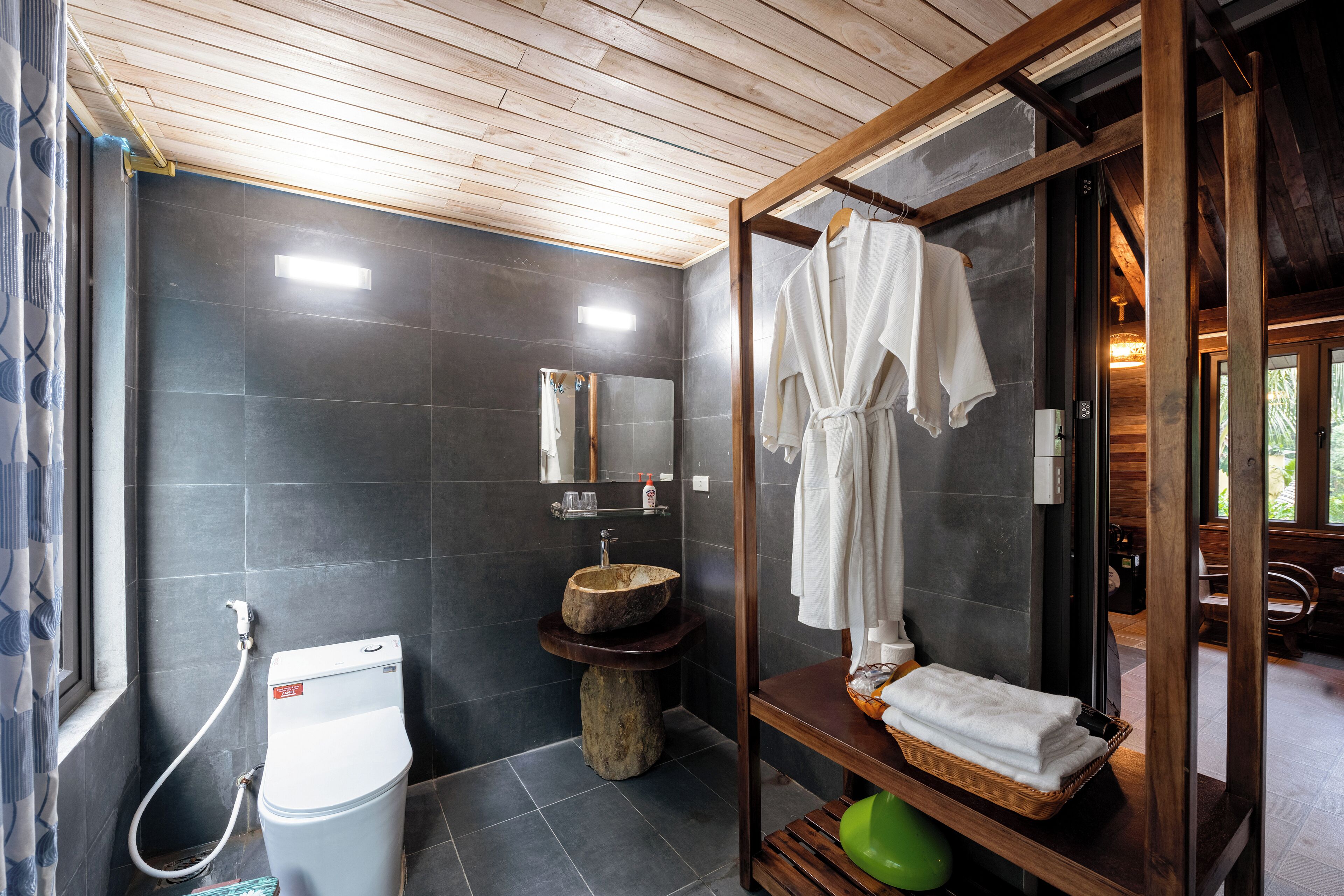 Un baño moderno con azulejos oscuros, un tocador de madera, cabina de ducha de vidrio y un inodoro a la vista.