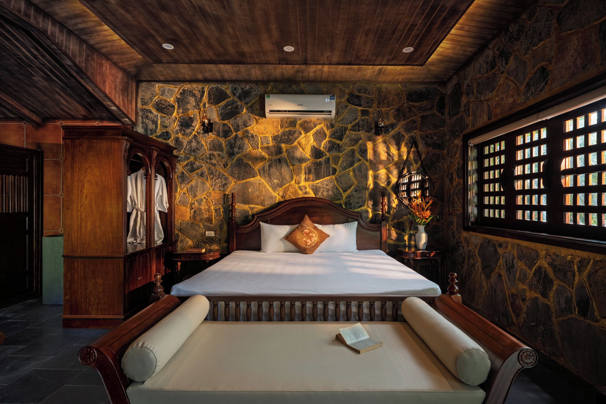 A bedroom with a stone wall, a bed with a wooden headboard, a nightstand, a wardrobe, and a window with a view.