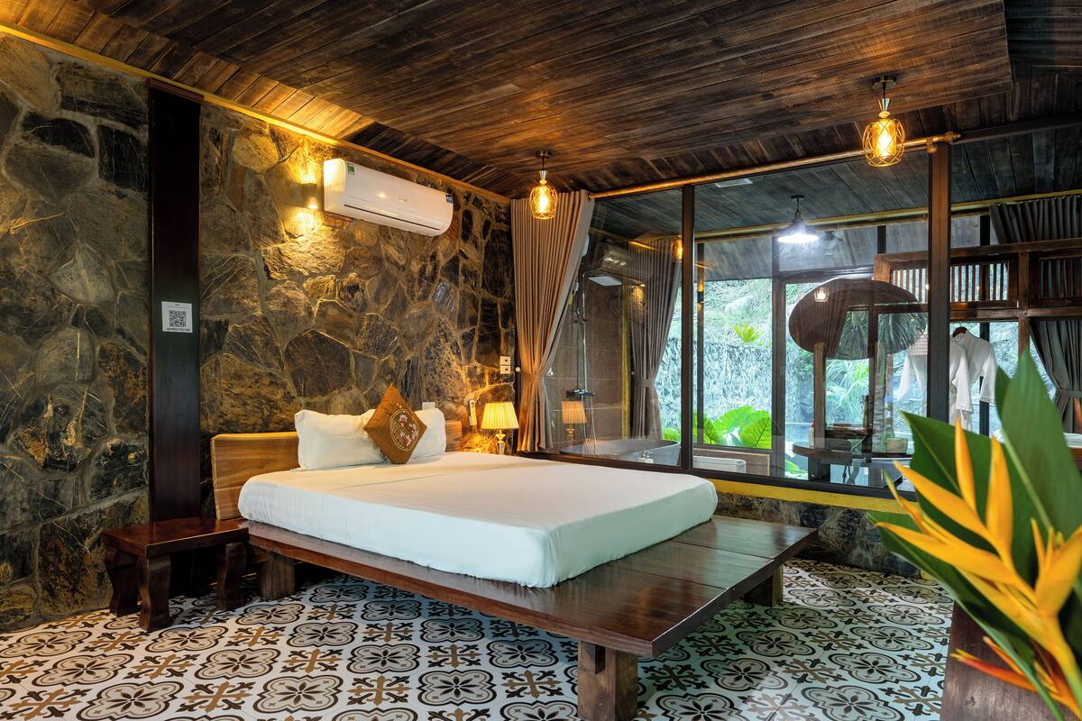 A bedroom with a stone wall, a wooden ceiling, a bed with white linens, a bedside table, and a window with curtains.