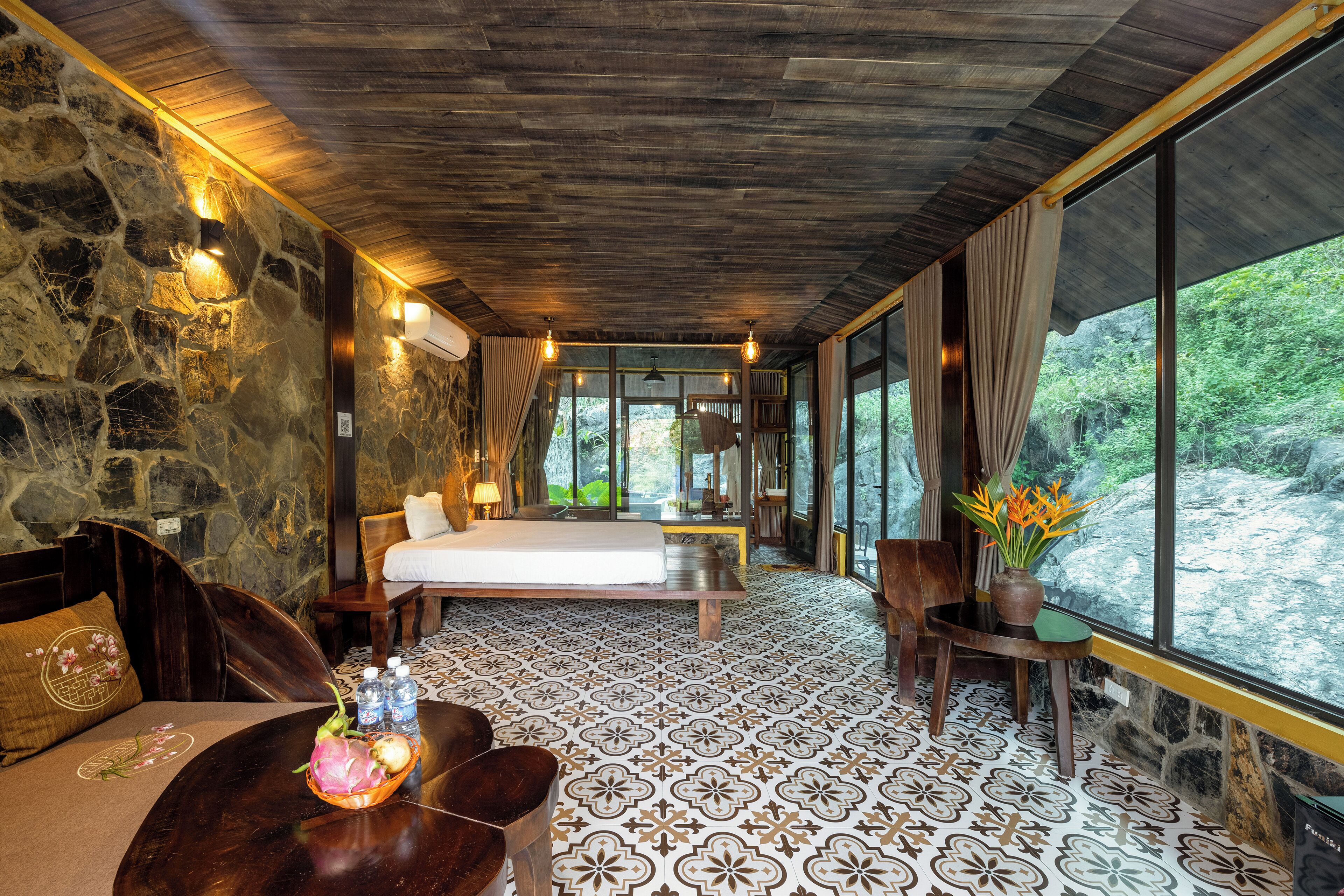 Une pièce avec un mur en pierre, un lit, une table à manger avec des chaises et une vue sur un paysage naturel, visible à travers de grandes fenêtres.