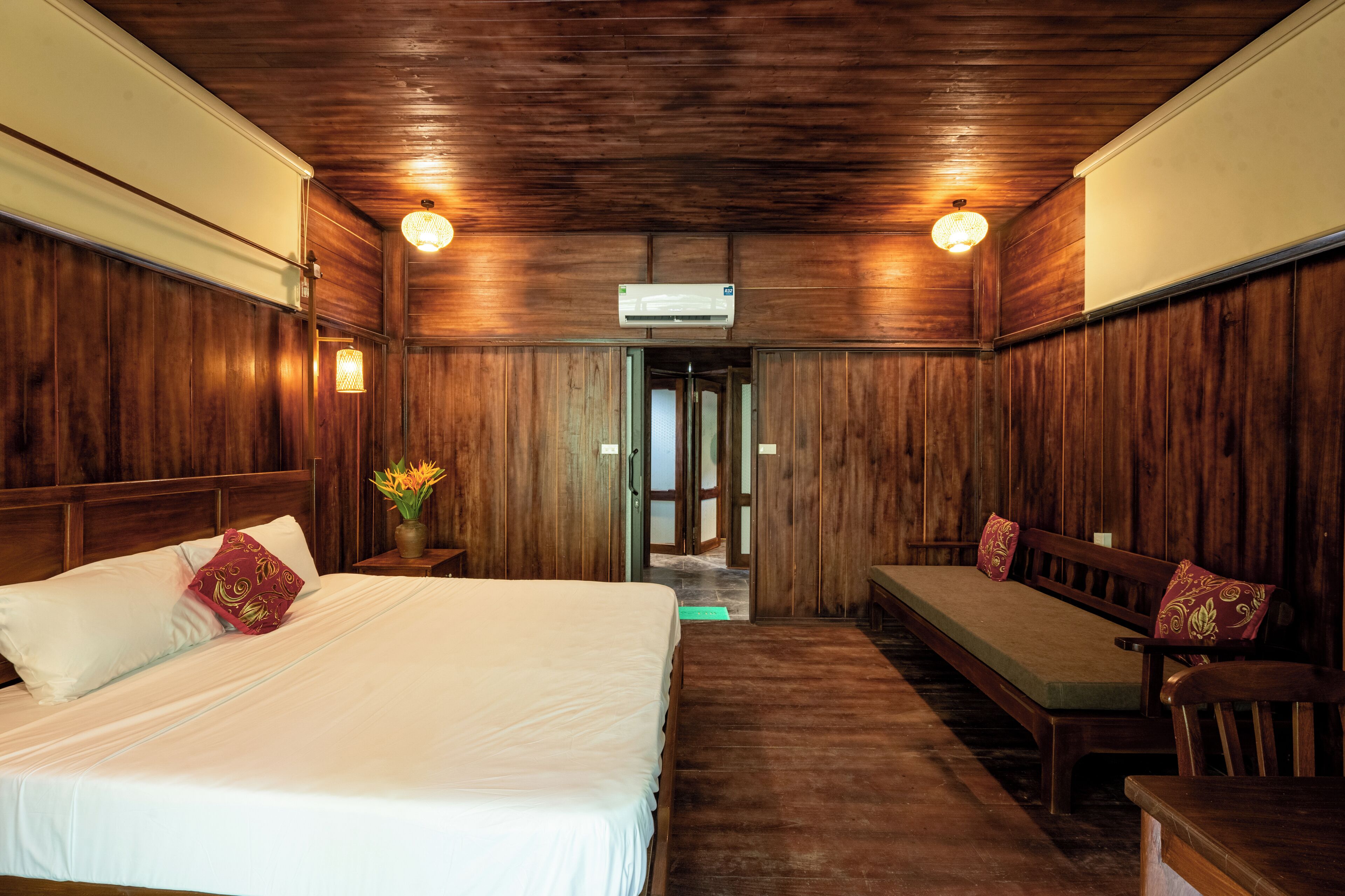 A room with a wooden ceiling, a bed with white bedding, a bench, and a small table with a vase of flowers.