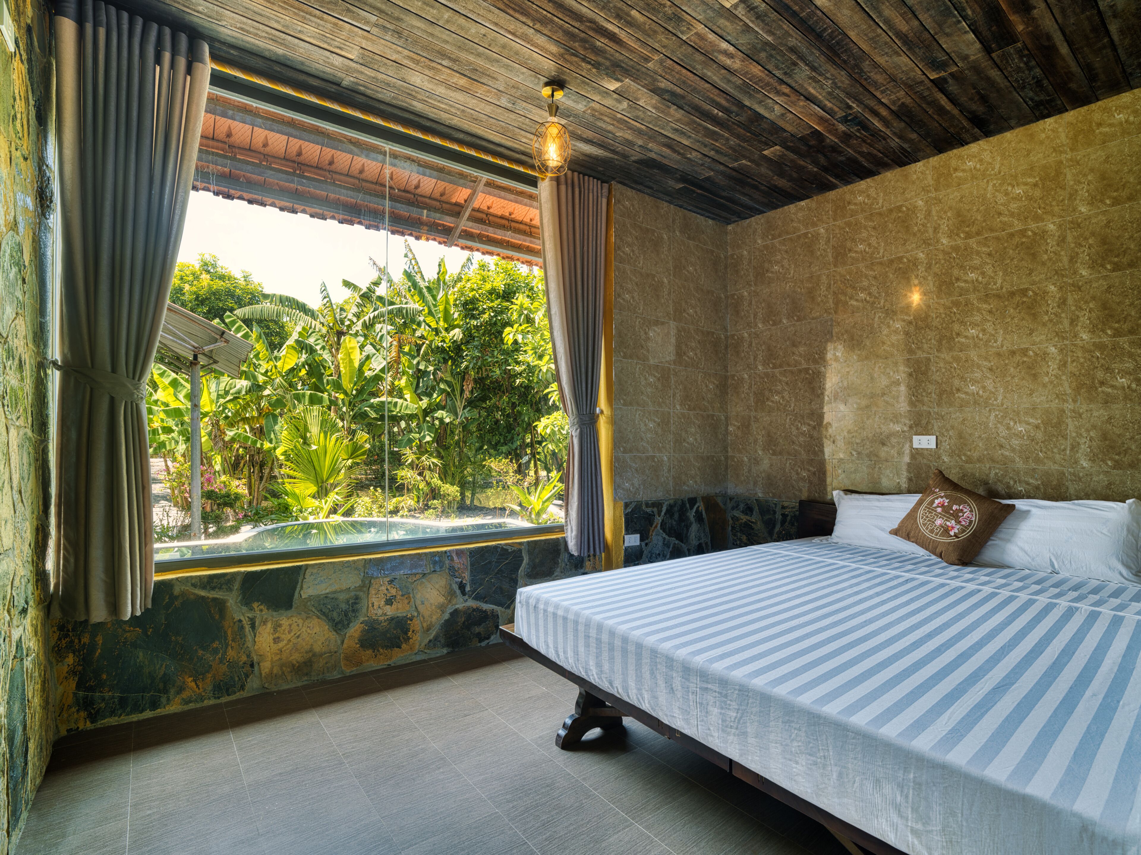 Un dormitorio con cama, cortinas, pared de piedra y vistas al verdor.