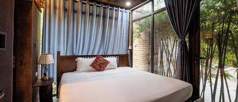 A bedroom with a large bed, a nightstand with a lamp, and a view of greenery through the window.