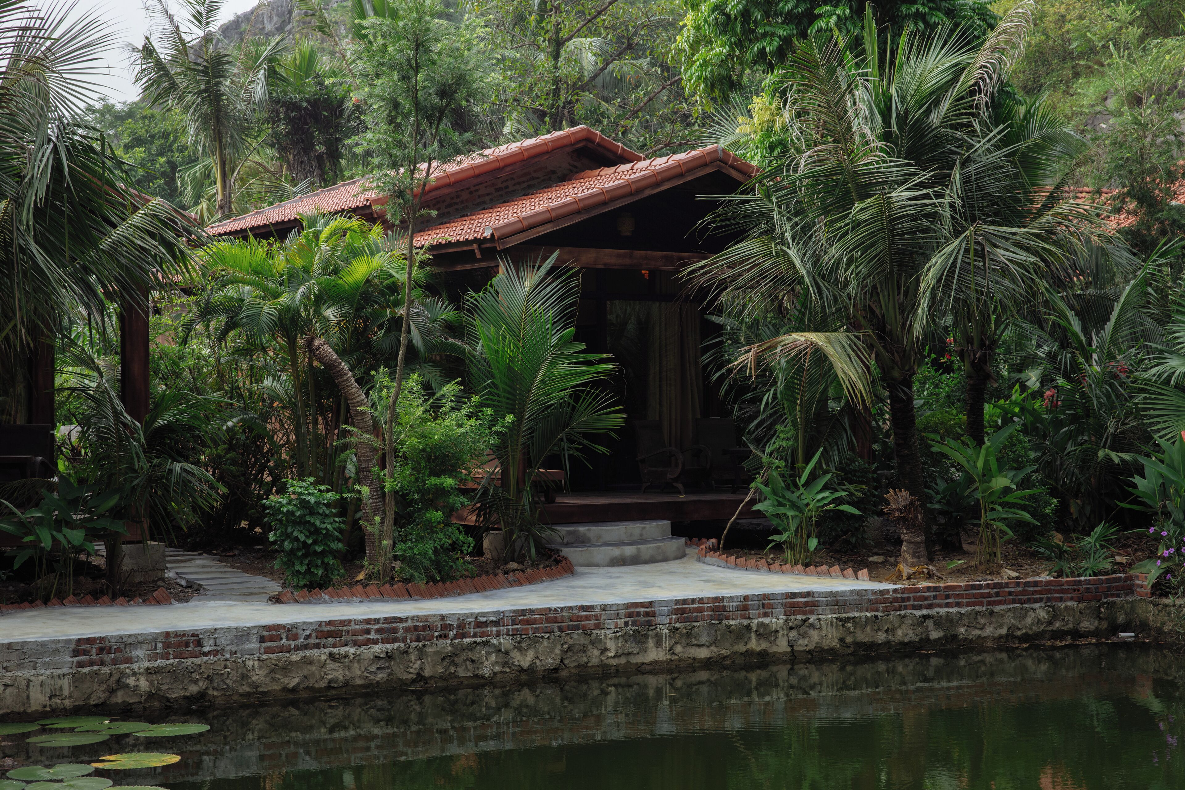 A traditional wooden structure with a tiled roof, surrounded by lush greenery and a calm water body.