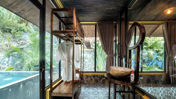 A room with a wooden bunk bed, a large window with curtains, a stone wall, and a view of a pool and greenery. - Trang An Garden Resort (Ninh Binh)