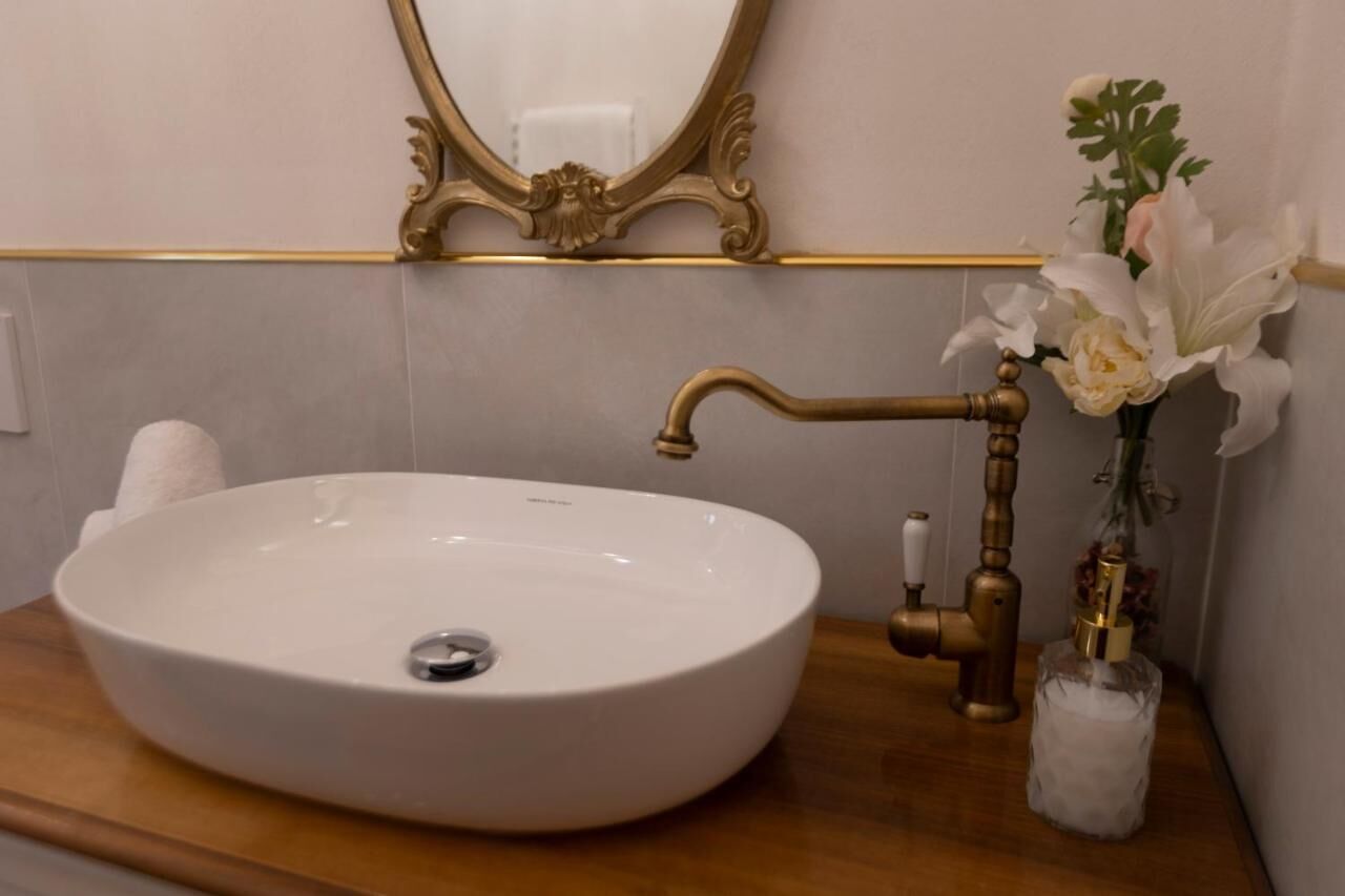 Un évier ovale blanc, un robinet en laiton sur un comptoir en bois, un miroir avec un cadre doré orné et un vase avec des fleurs blanches.