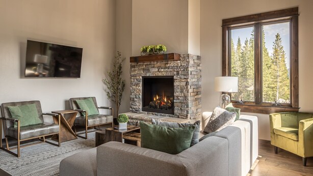A living room with a stone fireplace, two armchairs, a sofa, a TV mounted on the wall, and a window with a view of trees. - Condo with Private Hot Tub and Balcony Views | 5 Minutes to Ski Base (Big Sky)