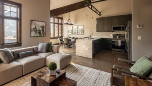 A modern living room with a sofa, coffee table, and a view of the kitchen. - Condo with Private Hot Tub and Balcony Views | 5 Minutes to Ski Base (Big Sky)