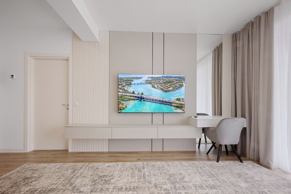 A modern living room with a large flat-screen TV mounted on the wall, a white desk, a grey chair, and a beige rug.