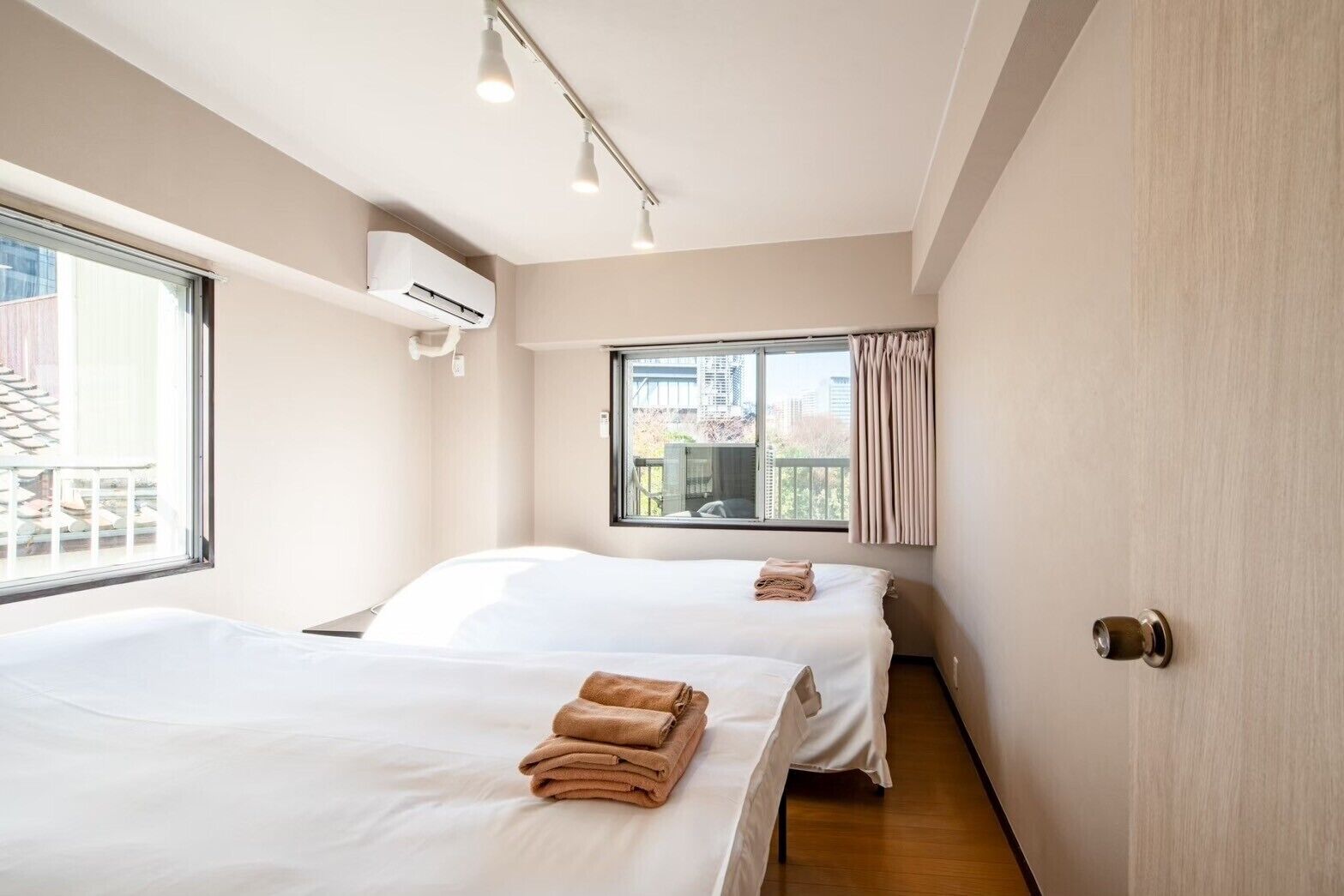 A hotel room with two beds, a window with curtains, an air conditioning unit, and a door with a handle.