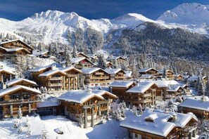Ein schneebedecktes Berghotel mit mehreren Holzhäusern, jedes mit Balkonen und großen Fenstern, umgeben von verschneiten Bäumen und Bergen.