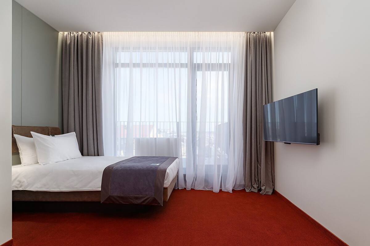 Hotellihuone, jossa on sänky, seinälle asennettu televisio ja läpikuultavat verhot.