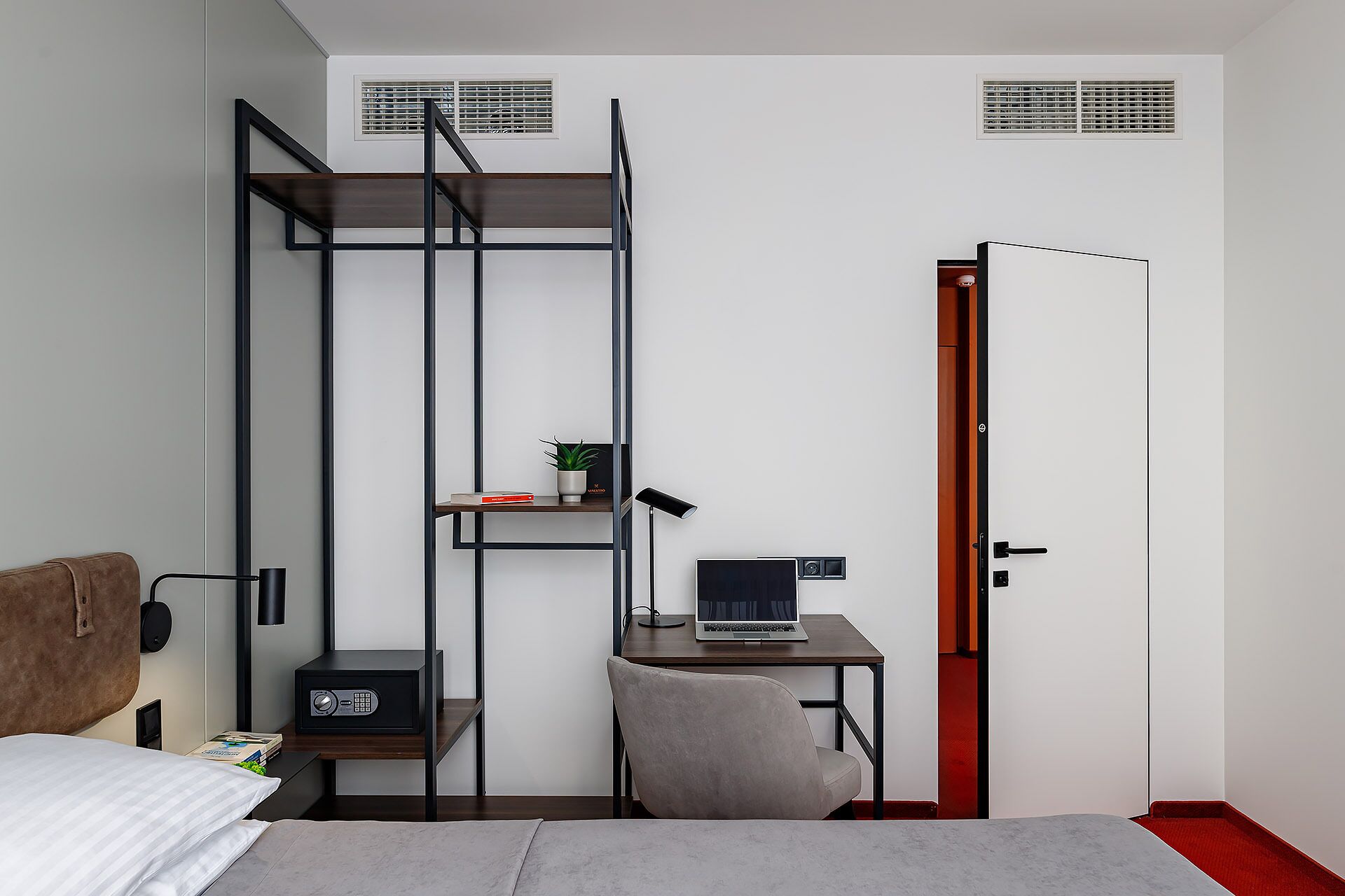 A compact bedroom with a bed, a desk with a laptop, a chair, a small shelf with a plant, and a radiator.