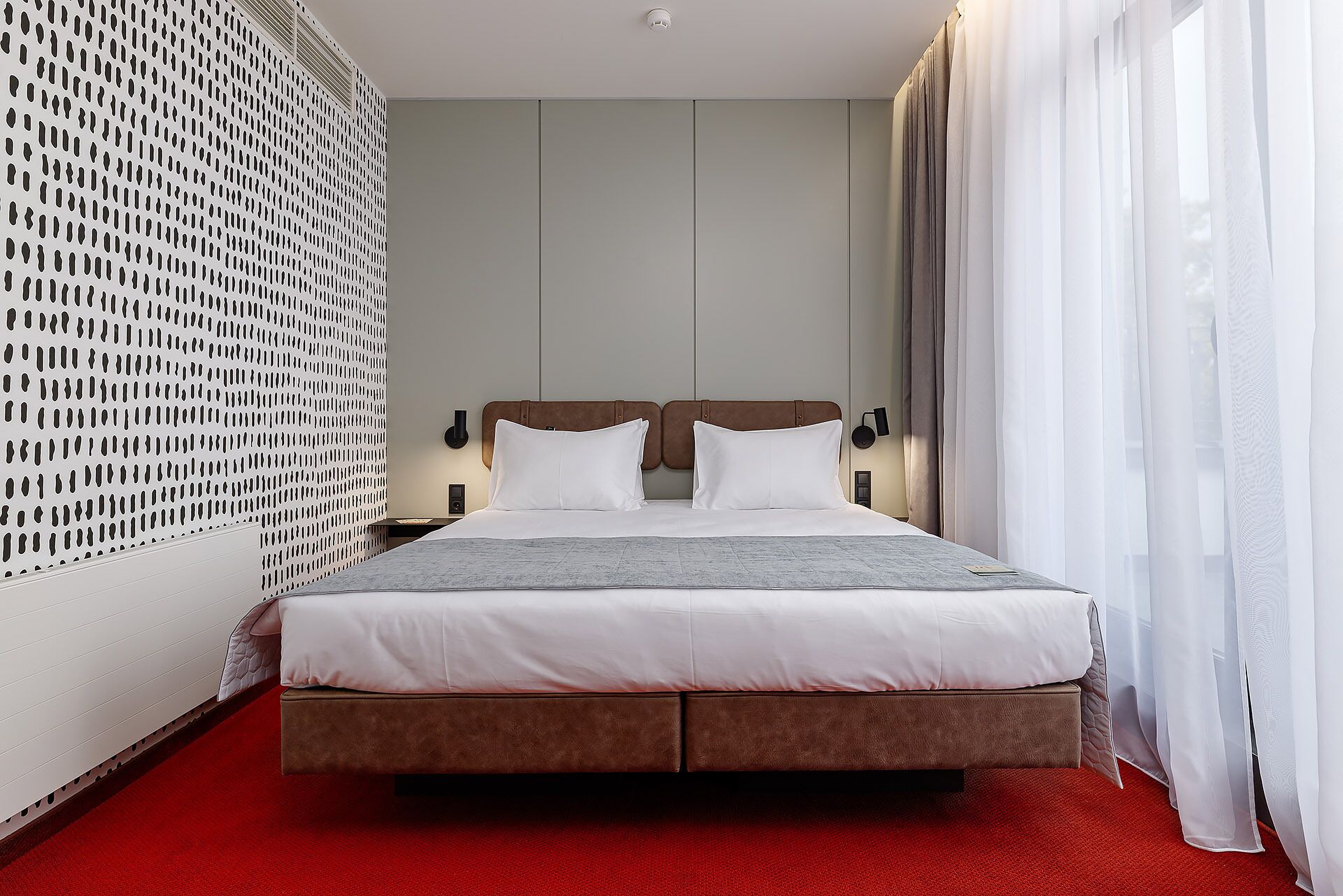 A hotel room with a large bed, a red carpet, a white wall with a perforated design, and a window with sheer curtains.