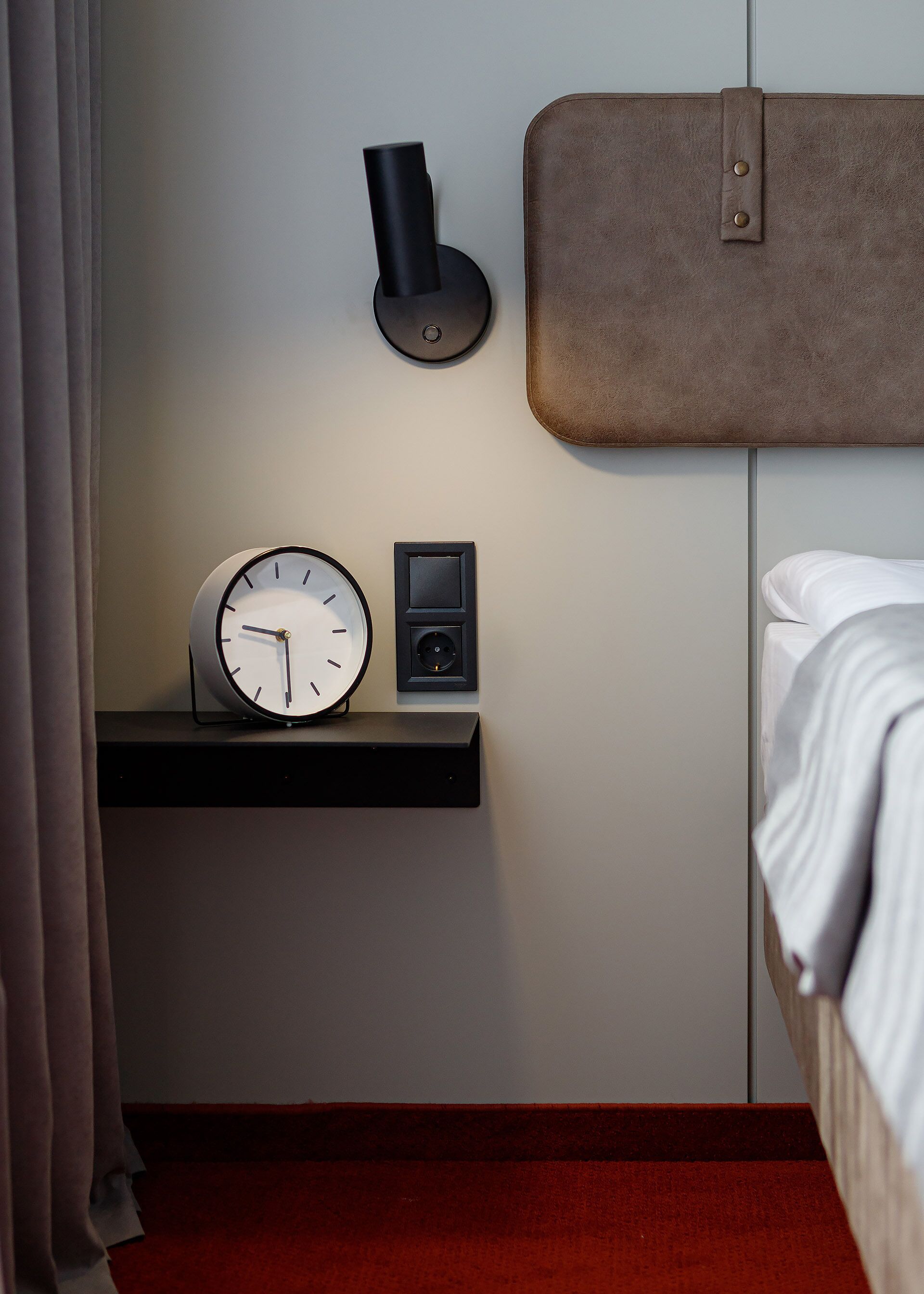 A hotel room with a bedside table featuring a wall clock, a small speaker, and a lamp.