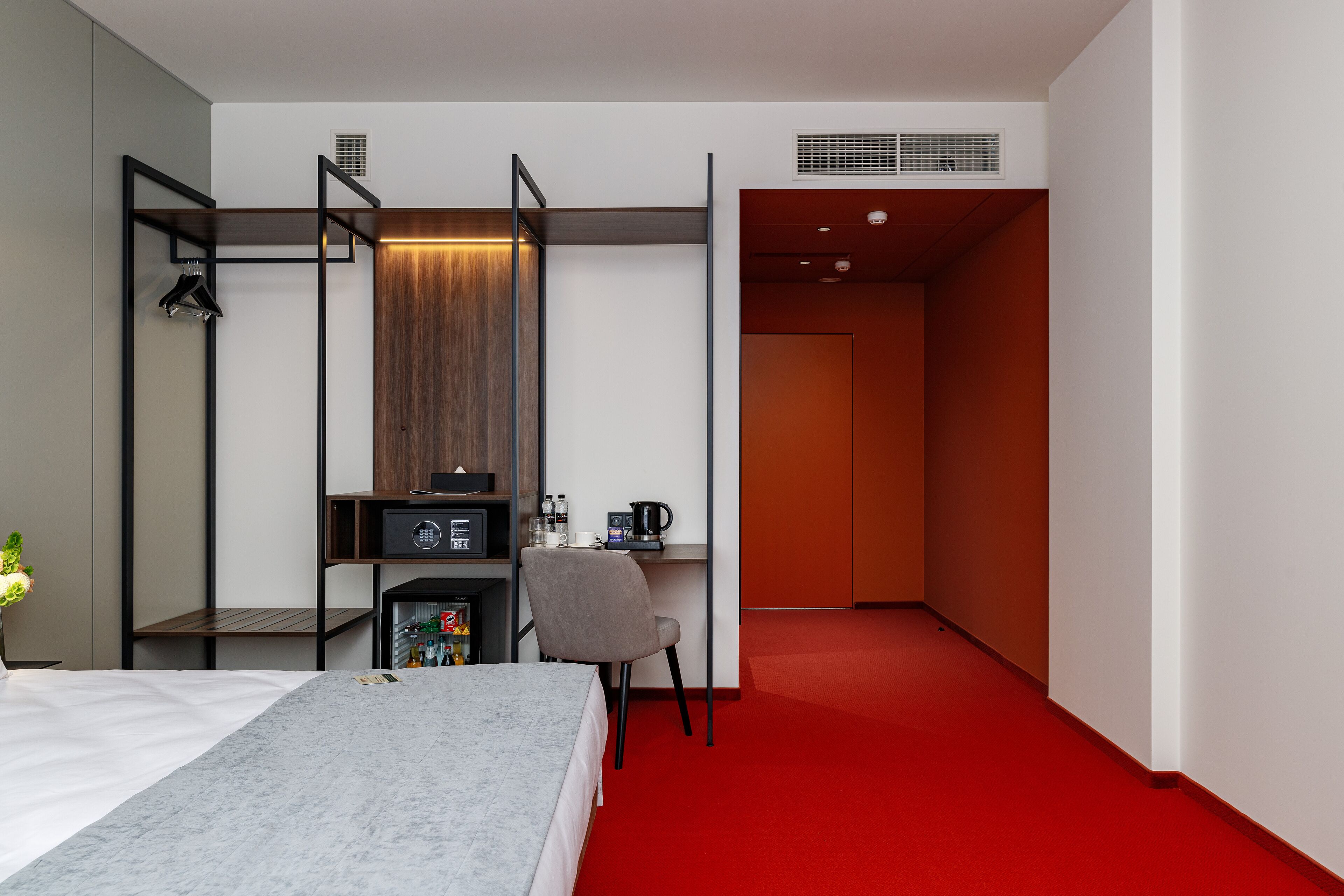 Une chambre d’hôtel comprenant un lit, un bureau avec une chaise, une armoire et un sol recouvert d’une moquette rouge.