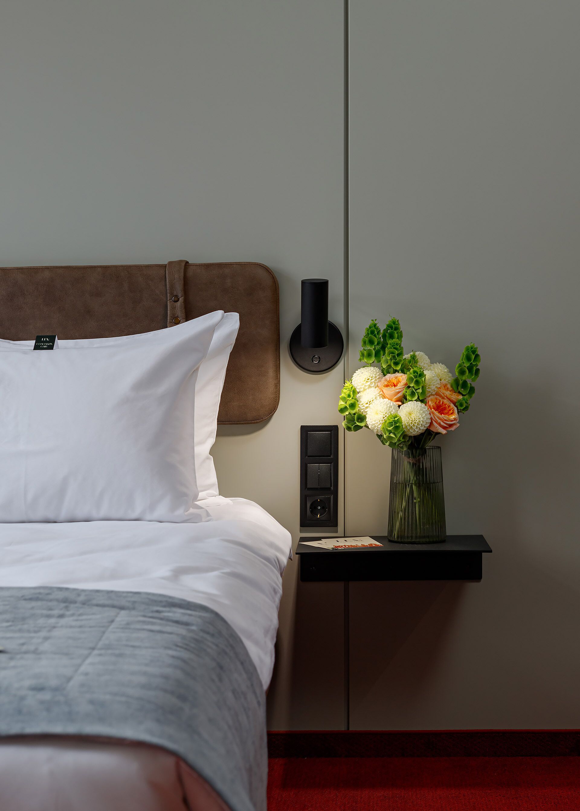 A neatly made bed with white linens, a brown headboard, a wall-mounted light fixture, a small nightstand with a vase of flowers, and a red carpeted floor.