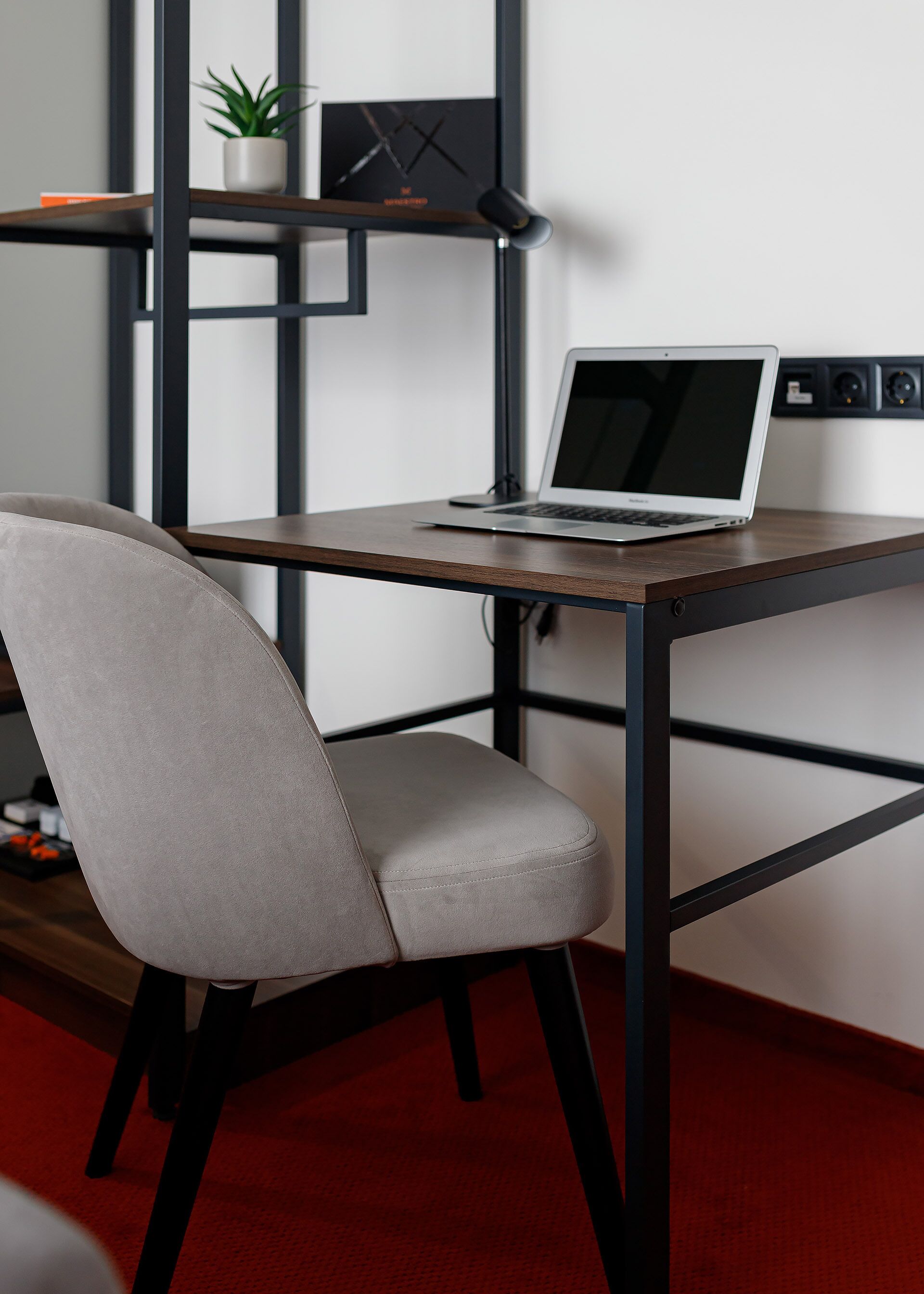 Una silla de oficina moderna con asiento y respaldo acolchados, un escritorio de madera con un ordenador portátil, y una estantería metálica negra con una planta en maceta.