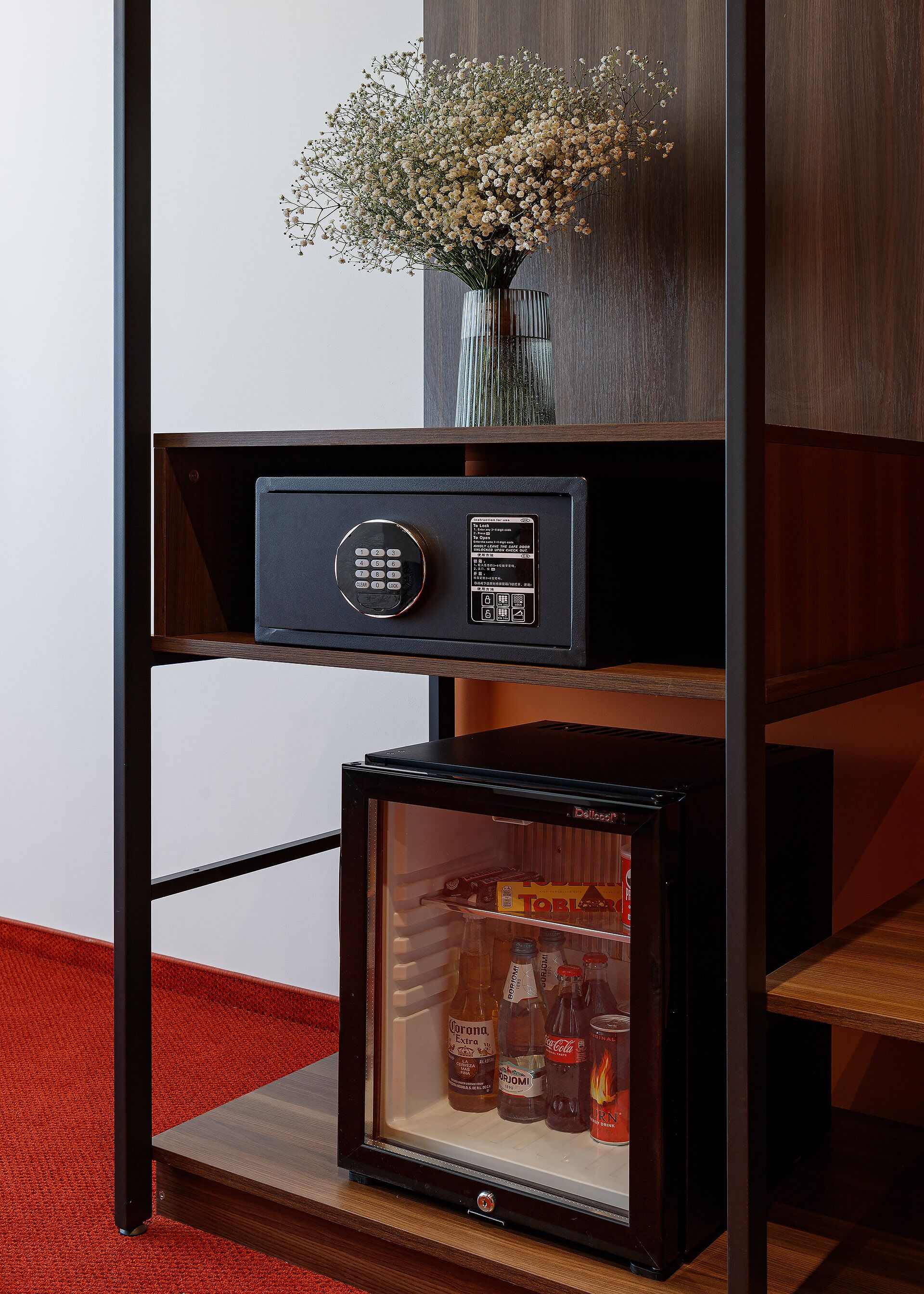 A secure storage unit with a keypad lock, a small refrigerator containing beverages, and a vase with flowers on top.