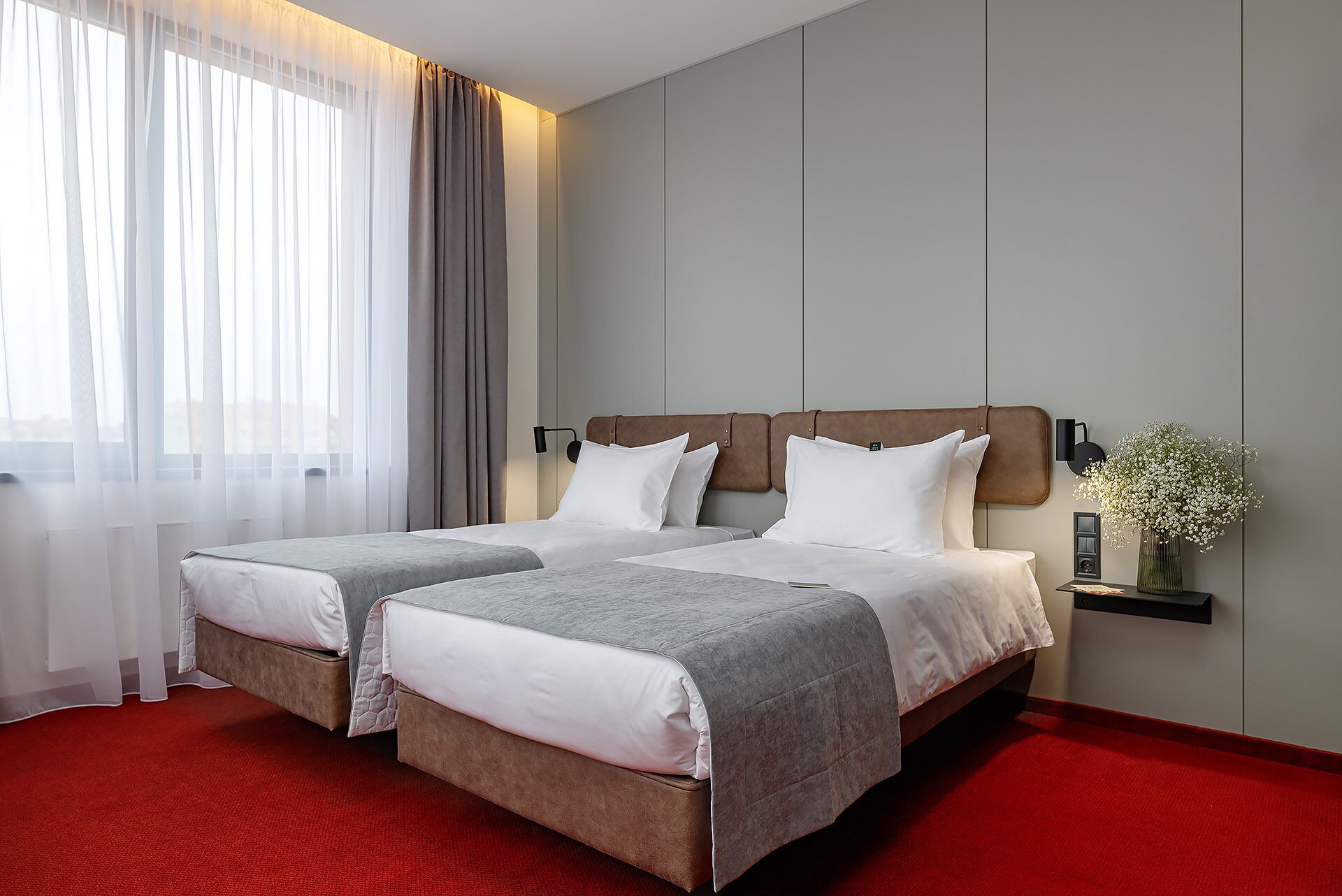A hotel room with two beds, a red carpet, a wall-mounted lamp, and a vase with flowers.