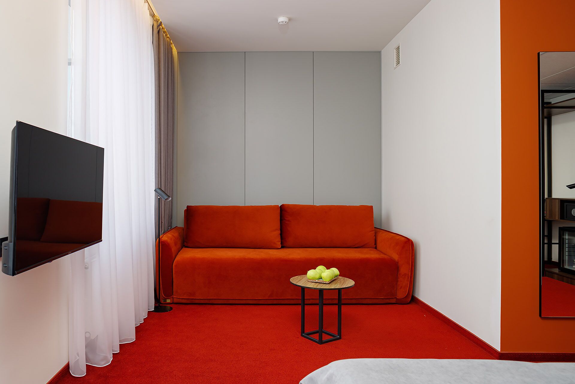 Habitación de hotel moderna con alfombra roja, un sofá con cojines naranjas, una pequeña mesa redonda con frutero, un televisor de pantalla plana en la pared y un espejo grande a la derecha.