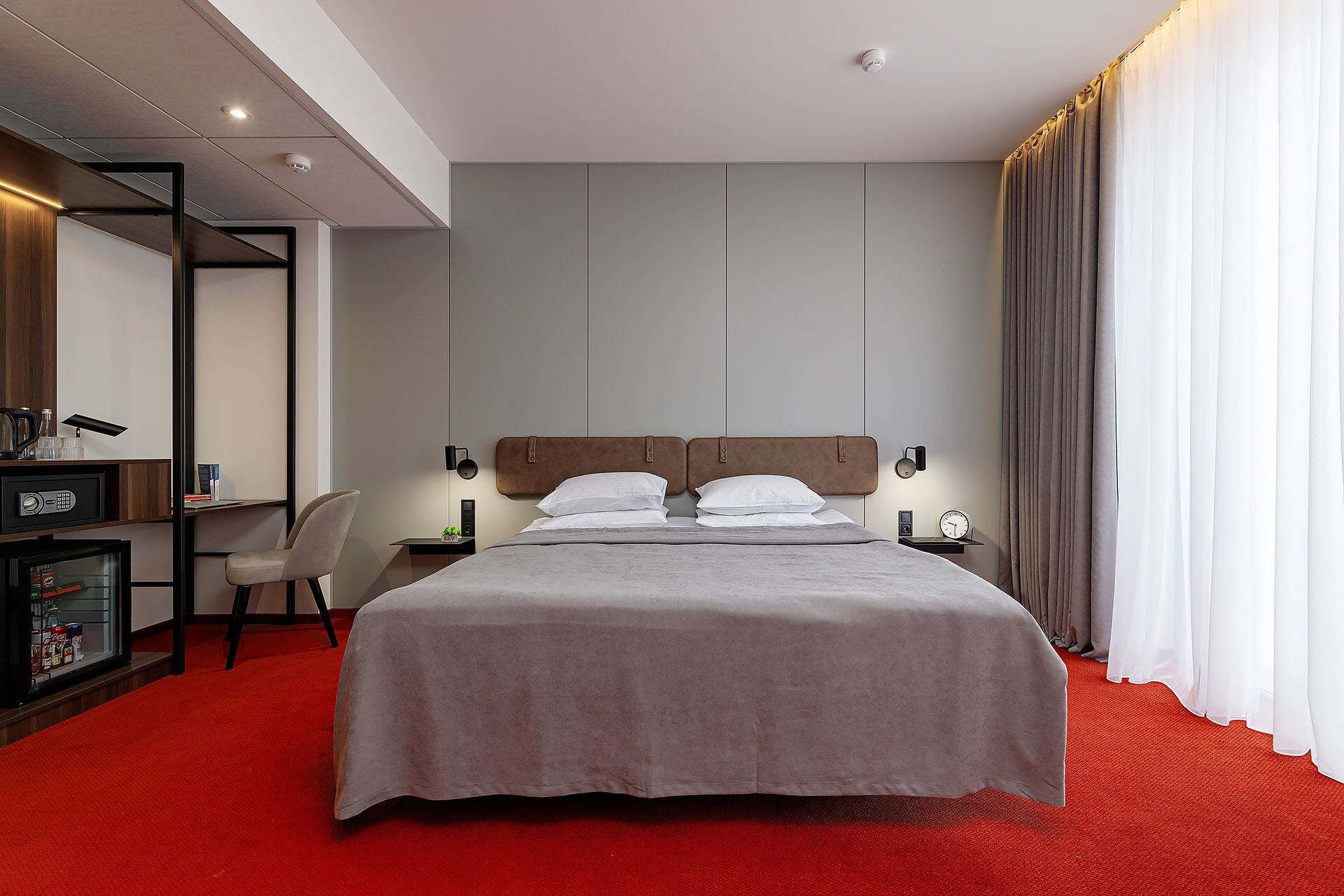 Een hotelkamer met een bed, een bureau met stoel, een kluisje en een rode tapijt.