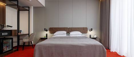 A hotel room with a bed, a desk with a chair, a safe, and a red carpet.
